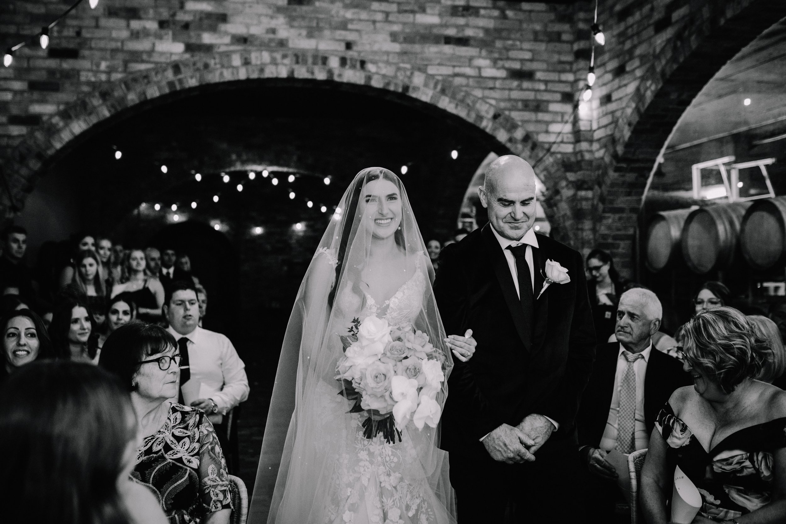 Black and white photo of a bride walking her father down the aisle during a wedding ceremony, with guests seated and smiling.
