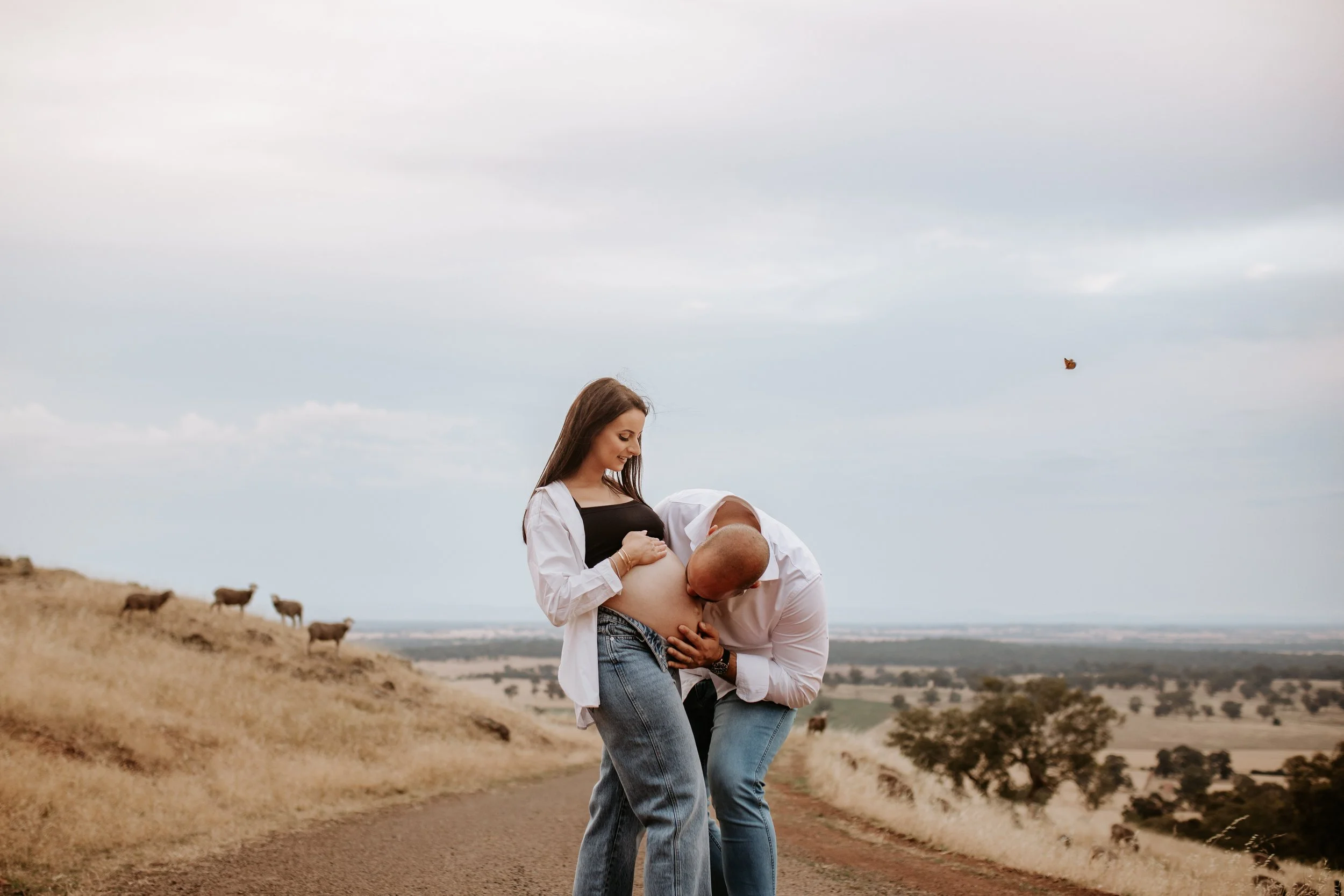 Pregnant woman with man kissing her belly in a rural outdoor landscape with cattle and open sky.