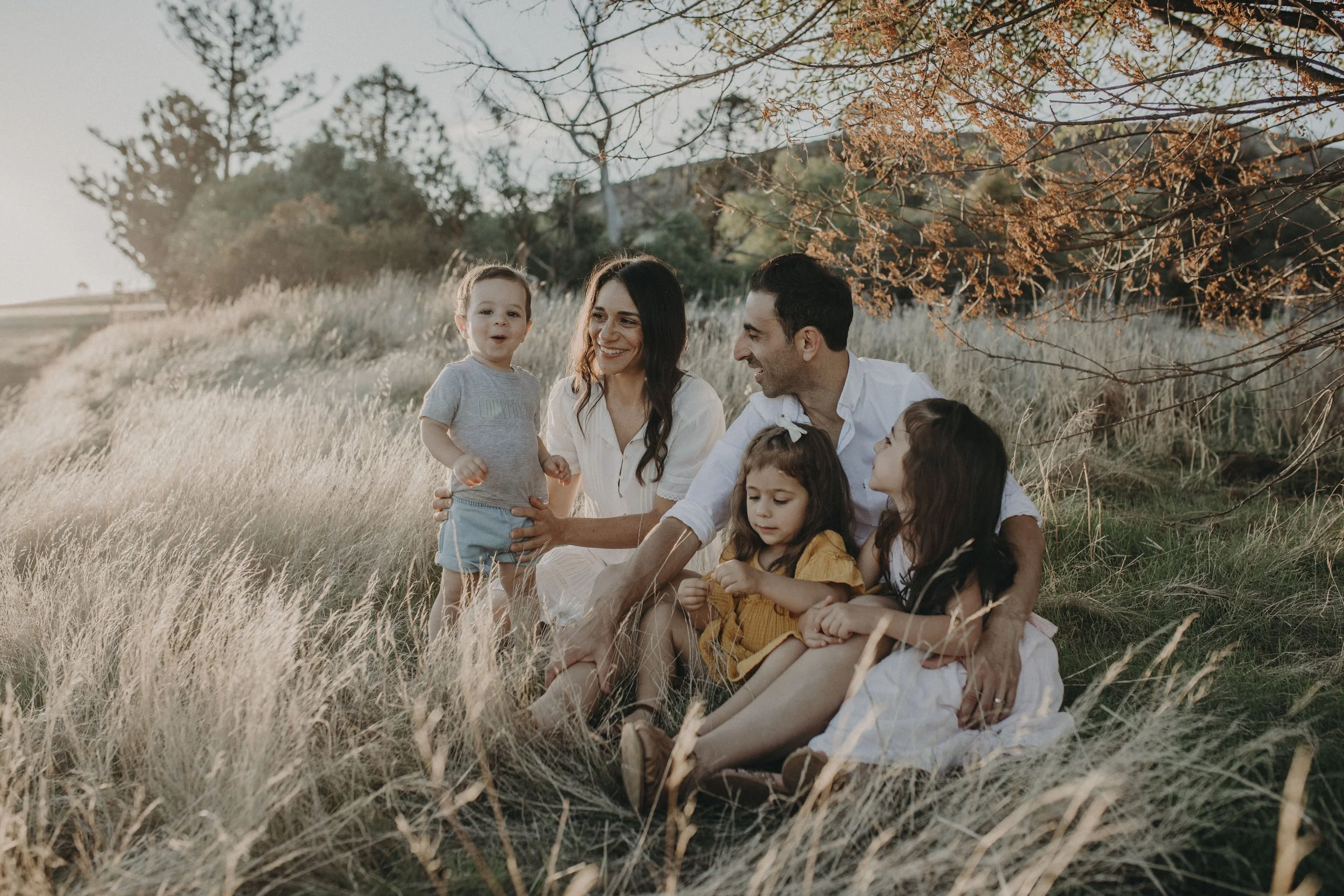 A family of five, including two adults and three children, sitting and playing in a grassy field with trees and autumn leaves in the background.