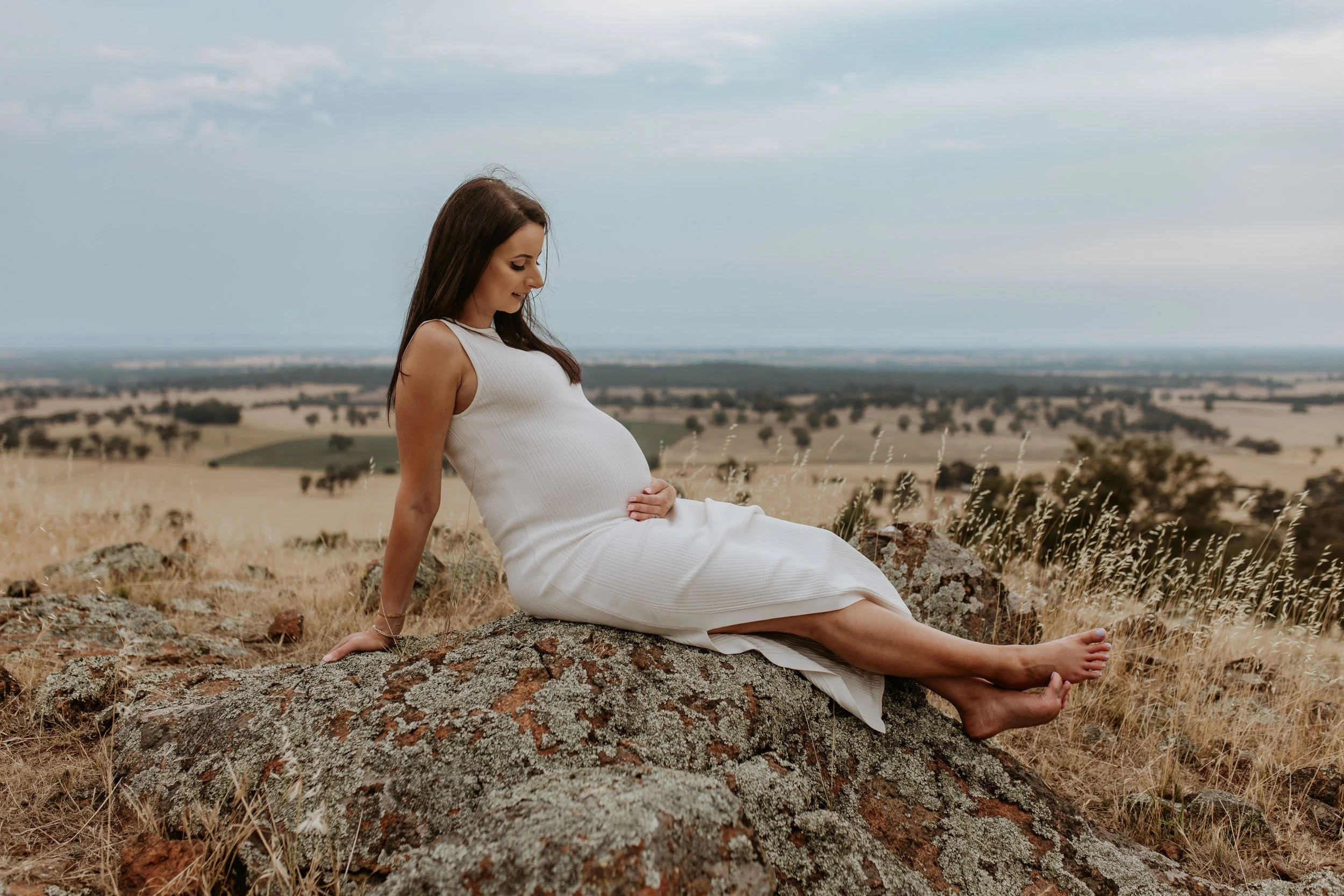 Pregnant woman in a white dress sitting on a rocky hilltop overlooking a rural landscape.