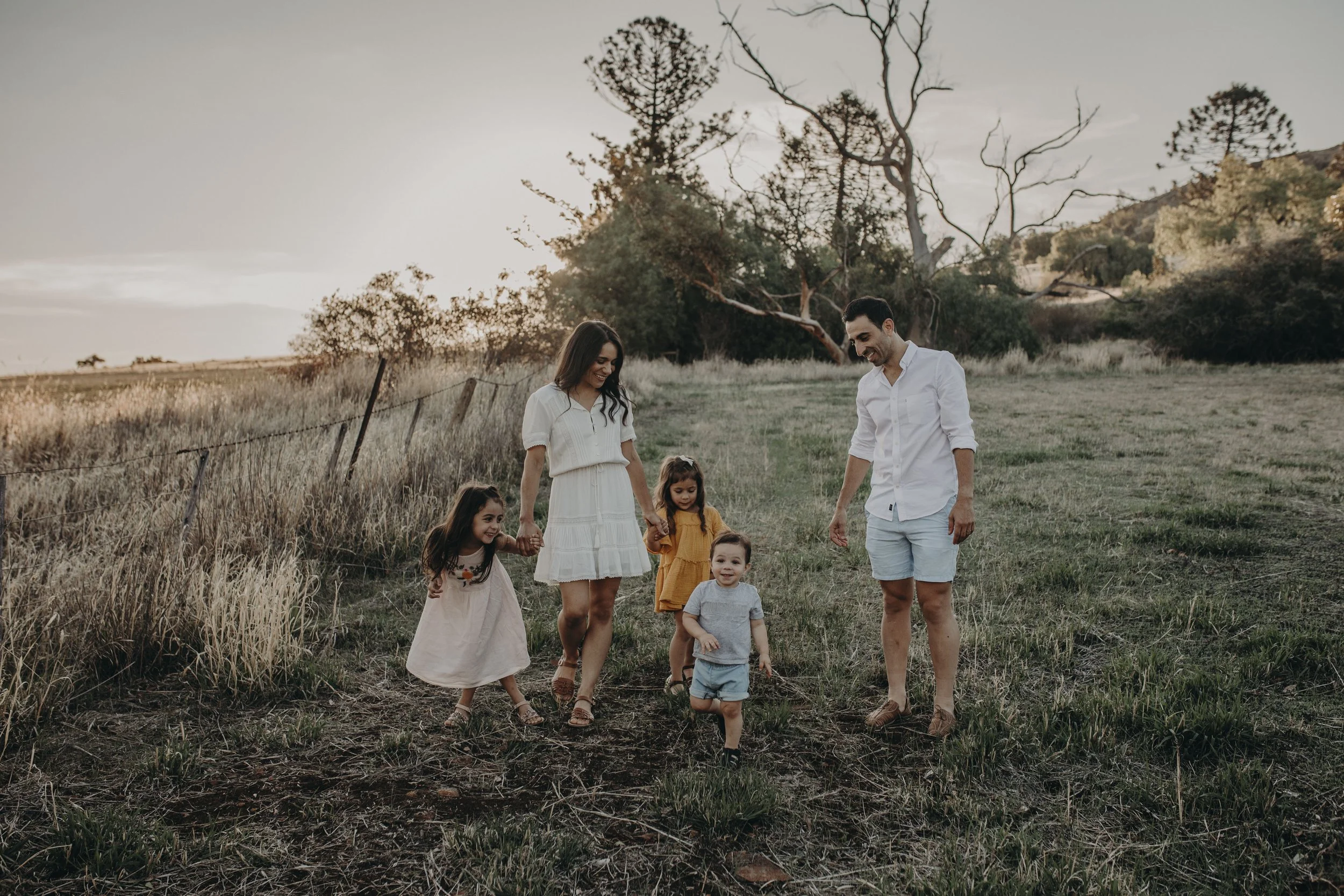 A family of five walking in a grassy field during sunset, holding hands, with trees in the background.