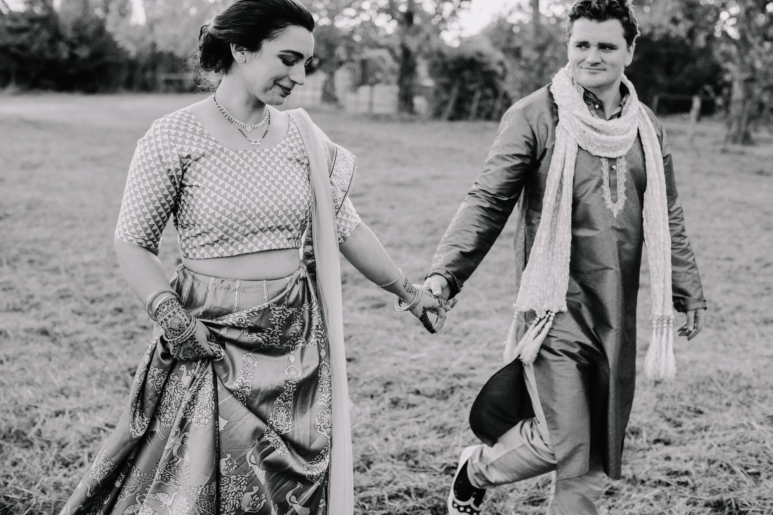 A man and woman dressed in traditional Indian attire, holding hands and walking outdoors in a grassy area, with trees in the background.