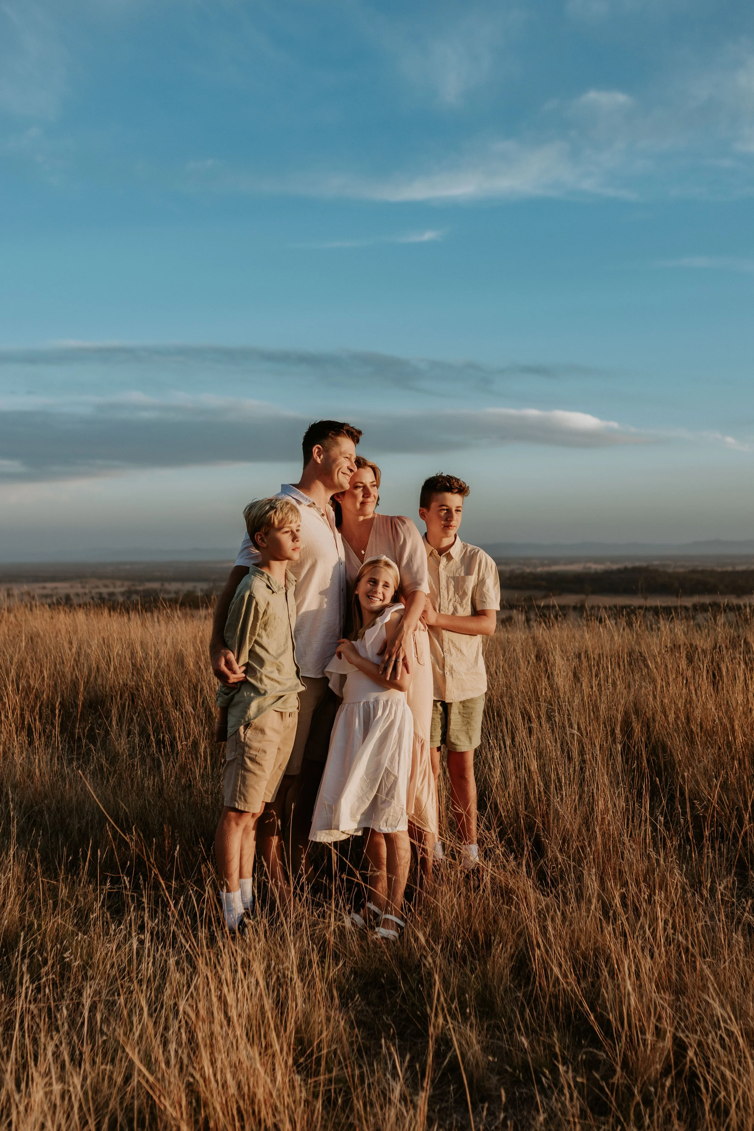 Family of five standing in a grassy field during sunset, embracing and smiling, with a sky filled with clouds in the background.