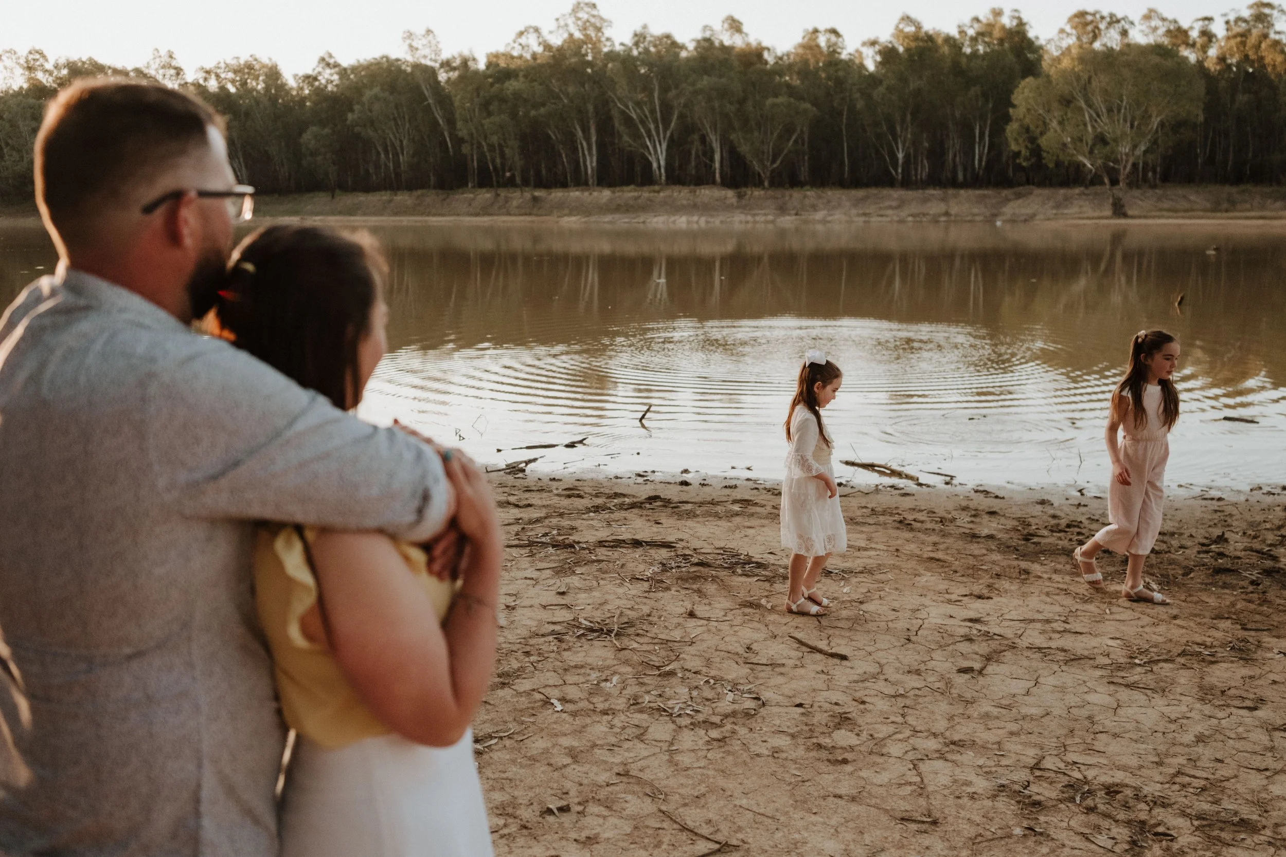 A family by a lake at sunset, including a man with glasses, a woman, and two small girls in white dresses, one holding the other as they walk near the water.