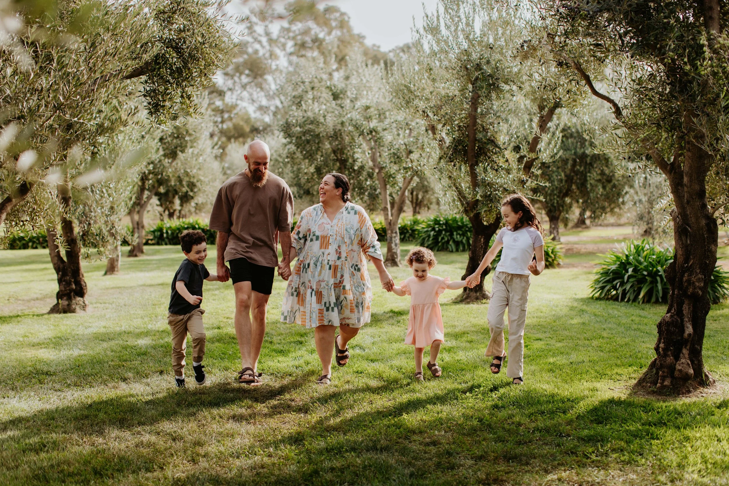 Family of five, including two adults and three children, walking hand-in-hand through a lush, green orchard with trees and sunlight overhead. Olivehouse, Kialla.