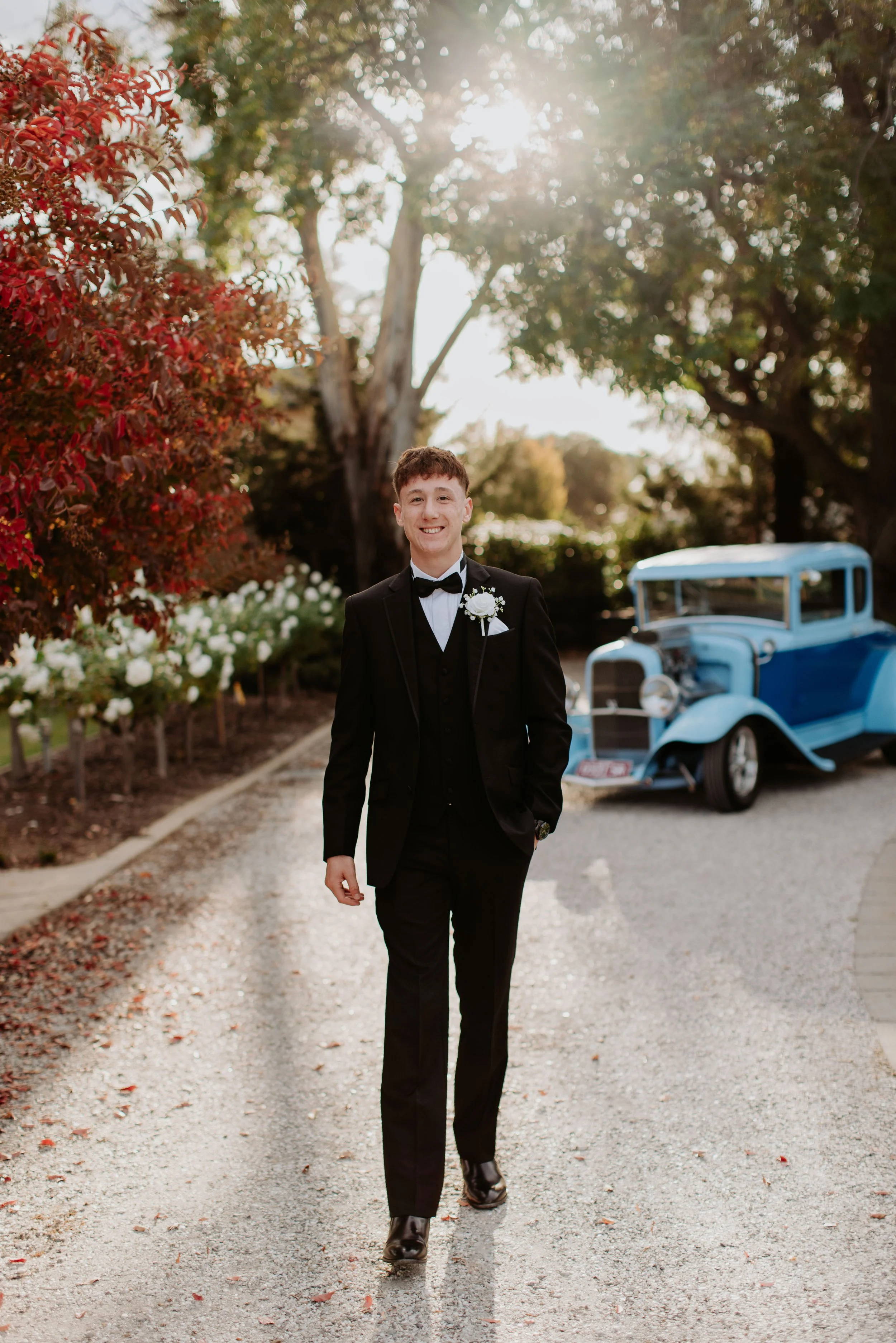 A young man in a tuxedo with a boutonniere walking on a gravel path, with a vintage blue car in the background and trees with autumn foliage.