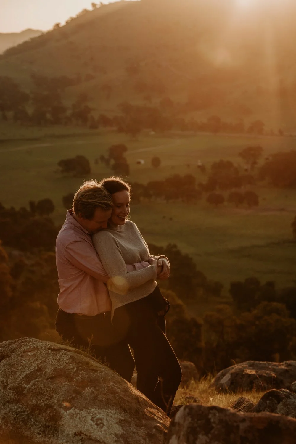 A happy couple hugging on a rocky hilltop with a sunset and rolling hills of Euroa in the background.