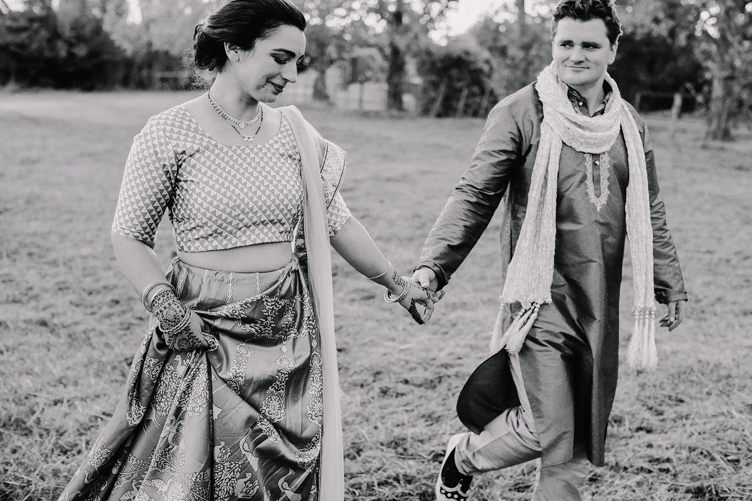 A man and woman dressed in traditional Indian attire holding hands outdoors, walking in a grassy field with trees in the background, in black and white.