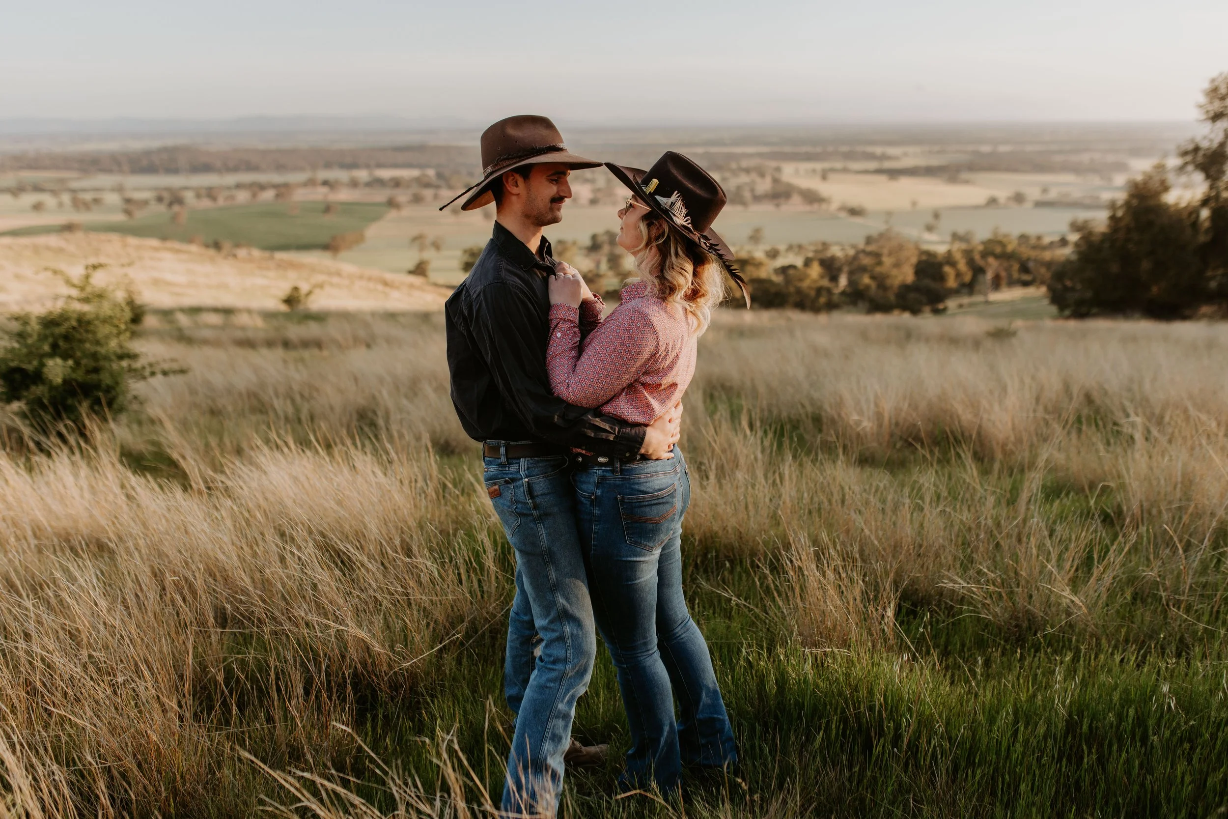 A couple wearing cowboy hats embracing in a grassy field during sunset with rolling hills in the background