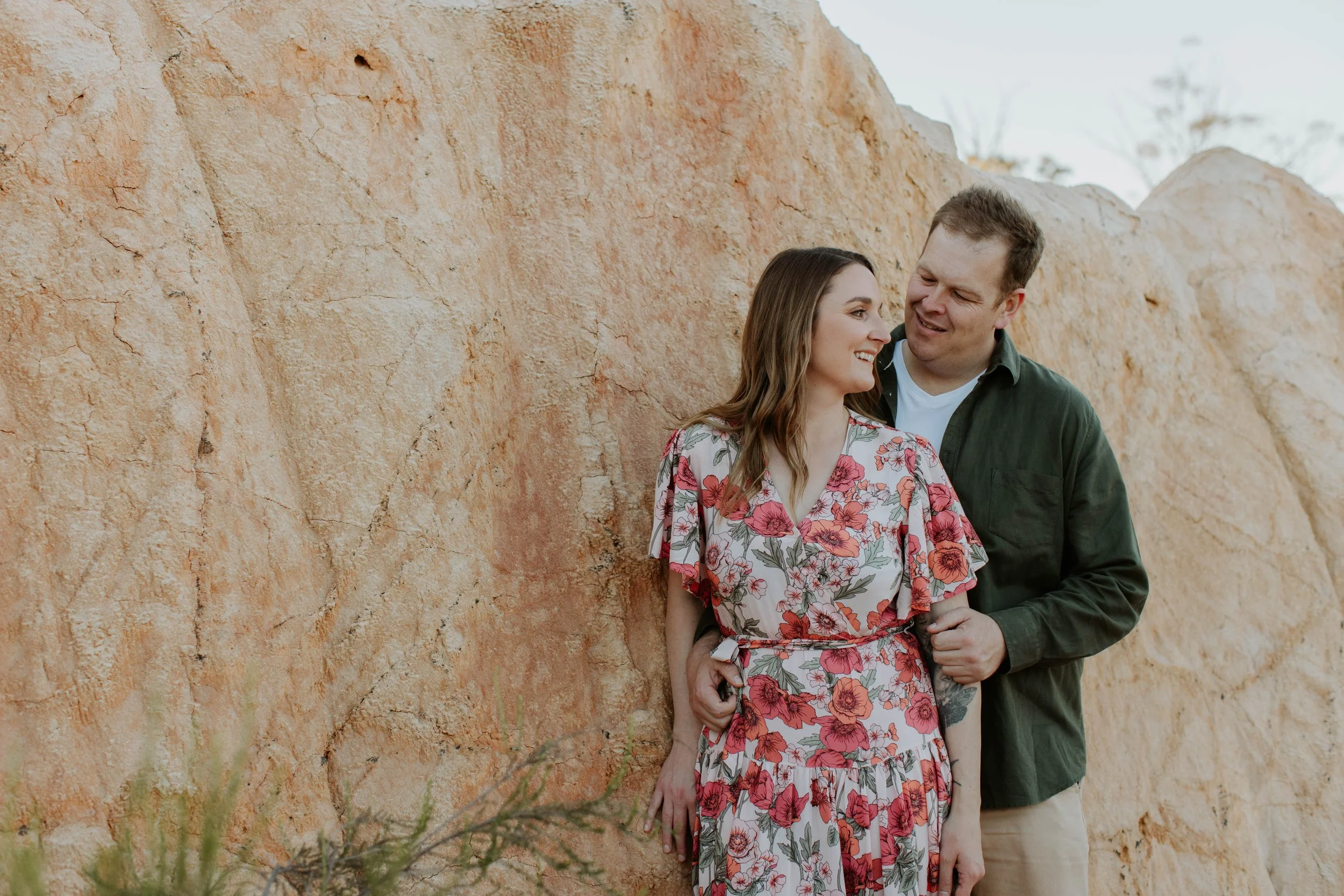 A smiling woman in a floral dress and a man in a green jacket stand close together outdoors in front of a large beige rock formation, sharing a moment of affection.