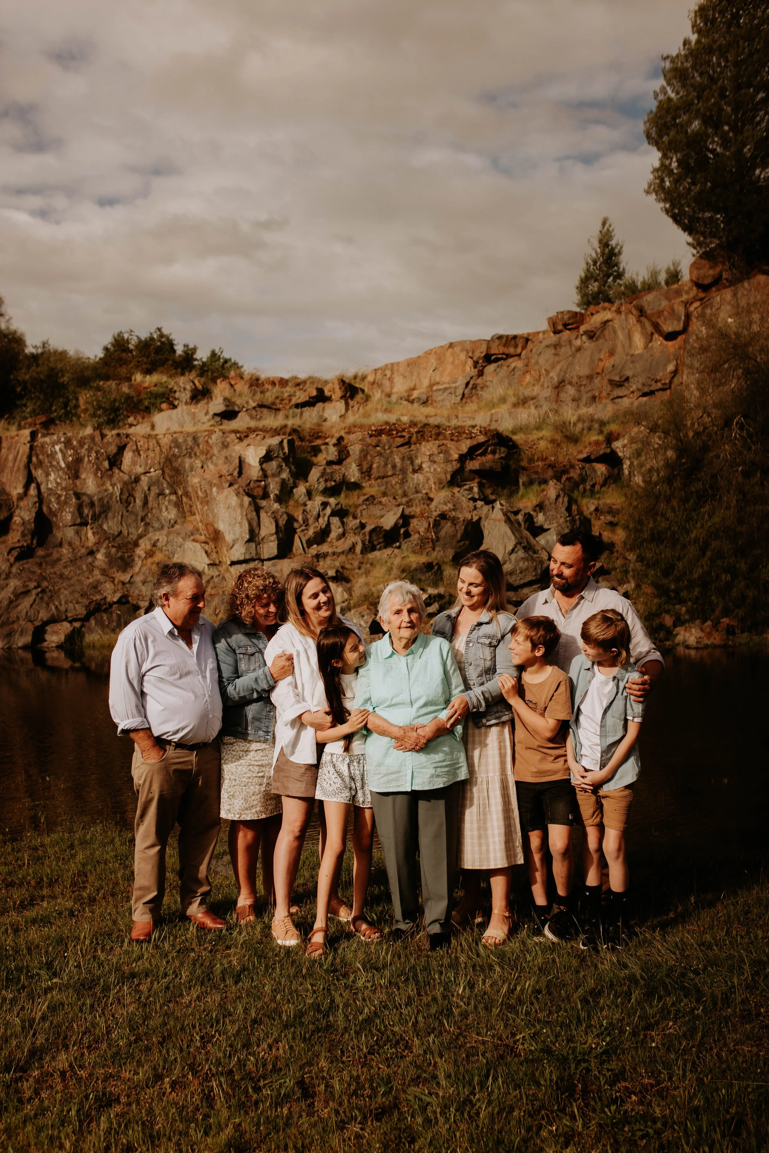 Family gathering in a natural outdoor setting near a river with rocky cliffs in the background, featuring grandparents, parents, and children smiling and embracing.