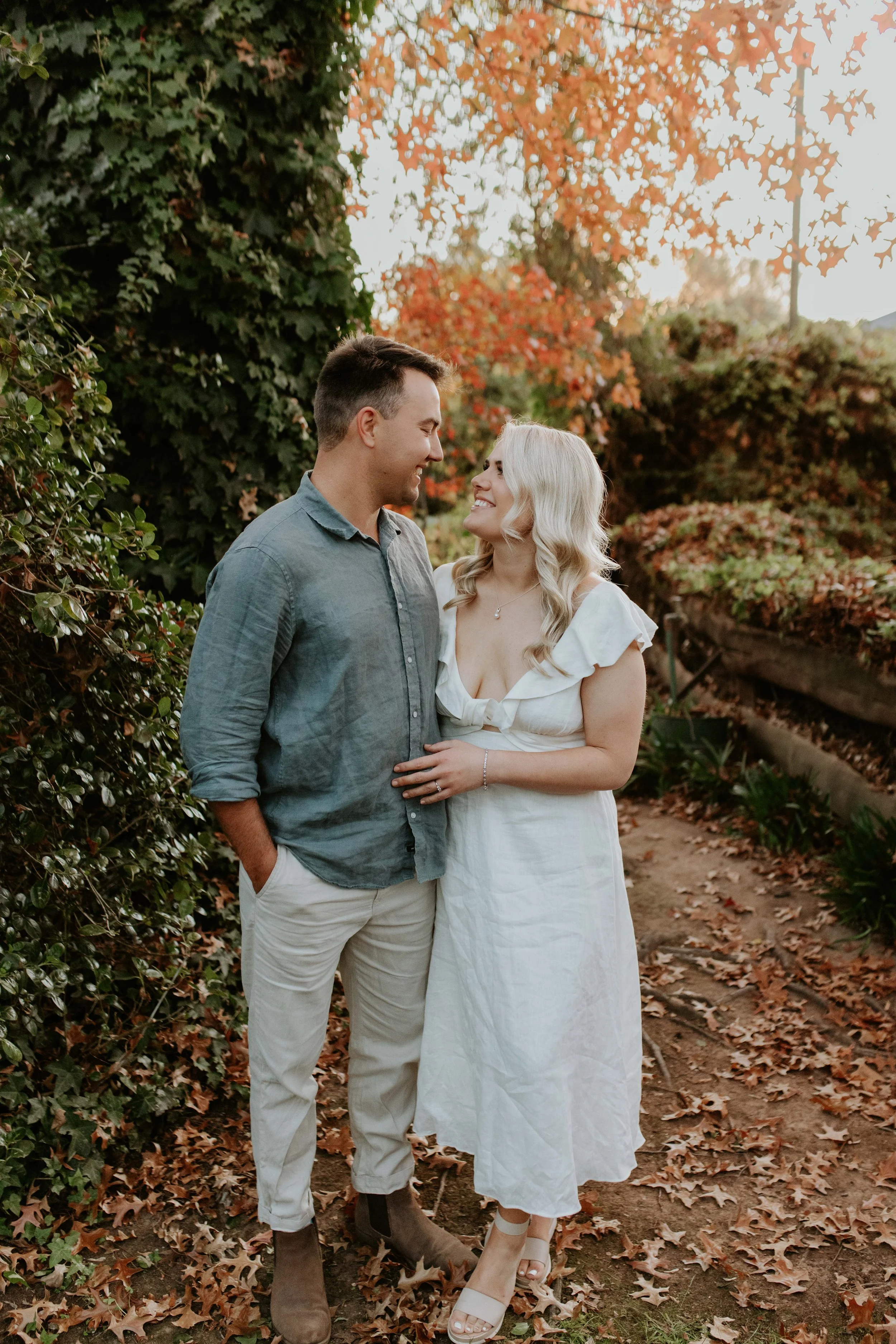 A couple standing close together outdoors among fall foliage, smiling and looking into each other's eyes.