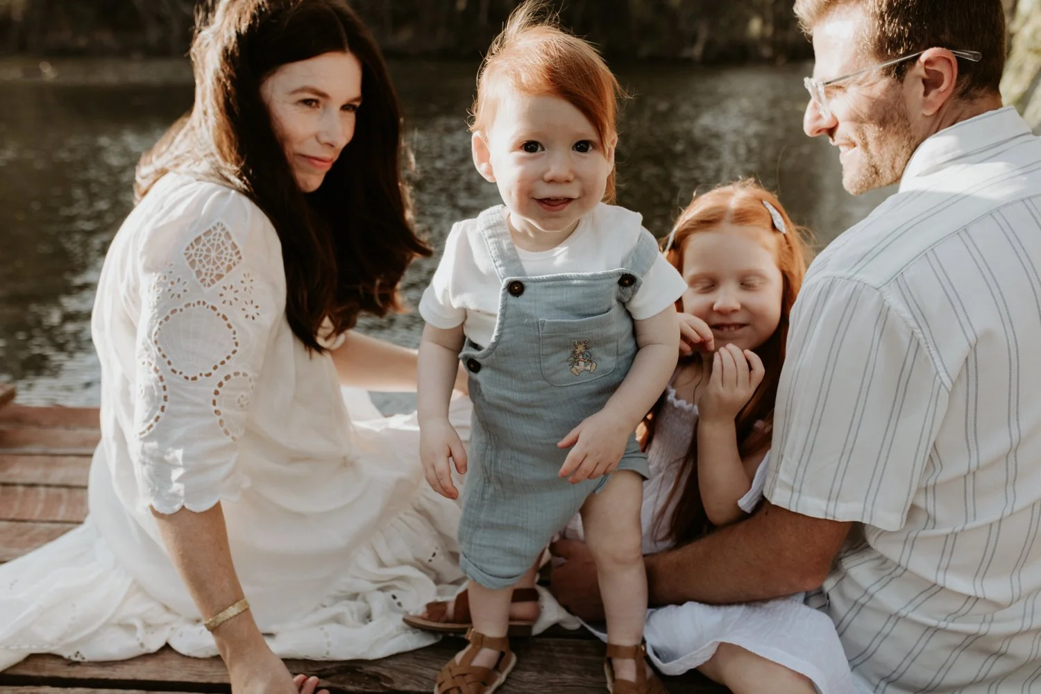 A family of four with a mother, father, and two young children enjoying time outdoors by a lake, sitting on a wooden dock during daytime. The children are redheaded, and everyone is smiling.