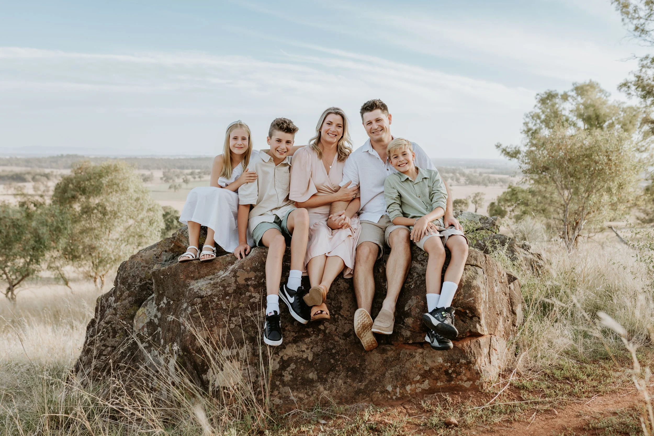 A family of five sitting on a large rock in a natural outdoor setting, smiling at the camera, with trees and open fields in the background. Mount Major, Dookie.