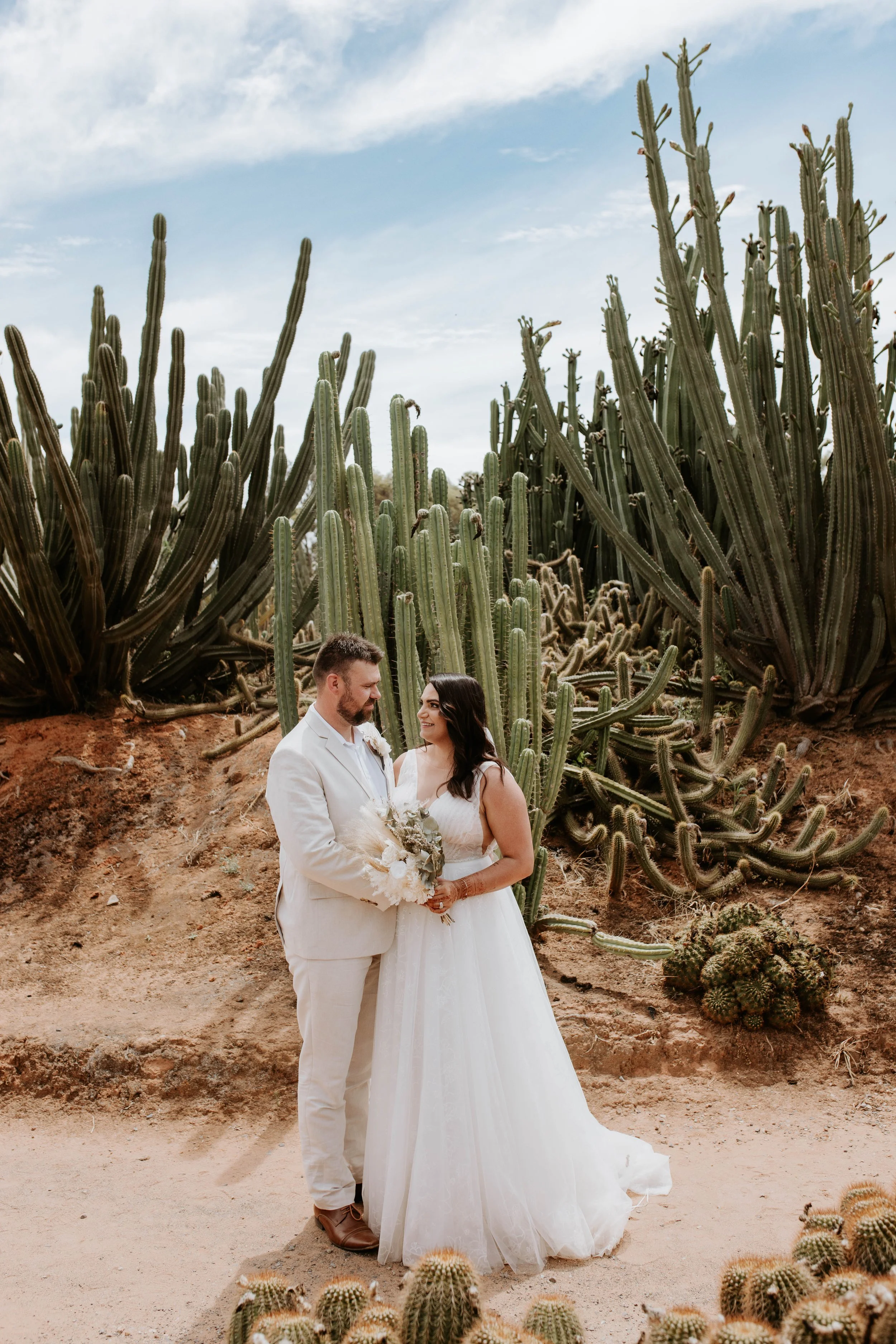 A bride and groom in wedding attire standing in front of large cacti in a desert setting.