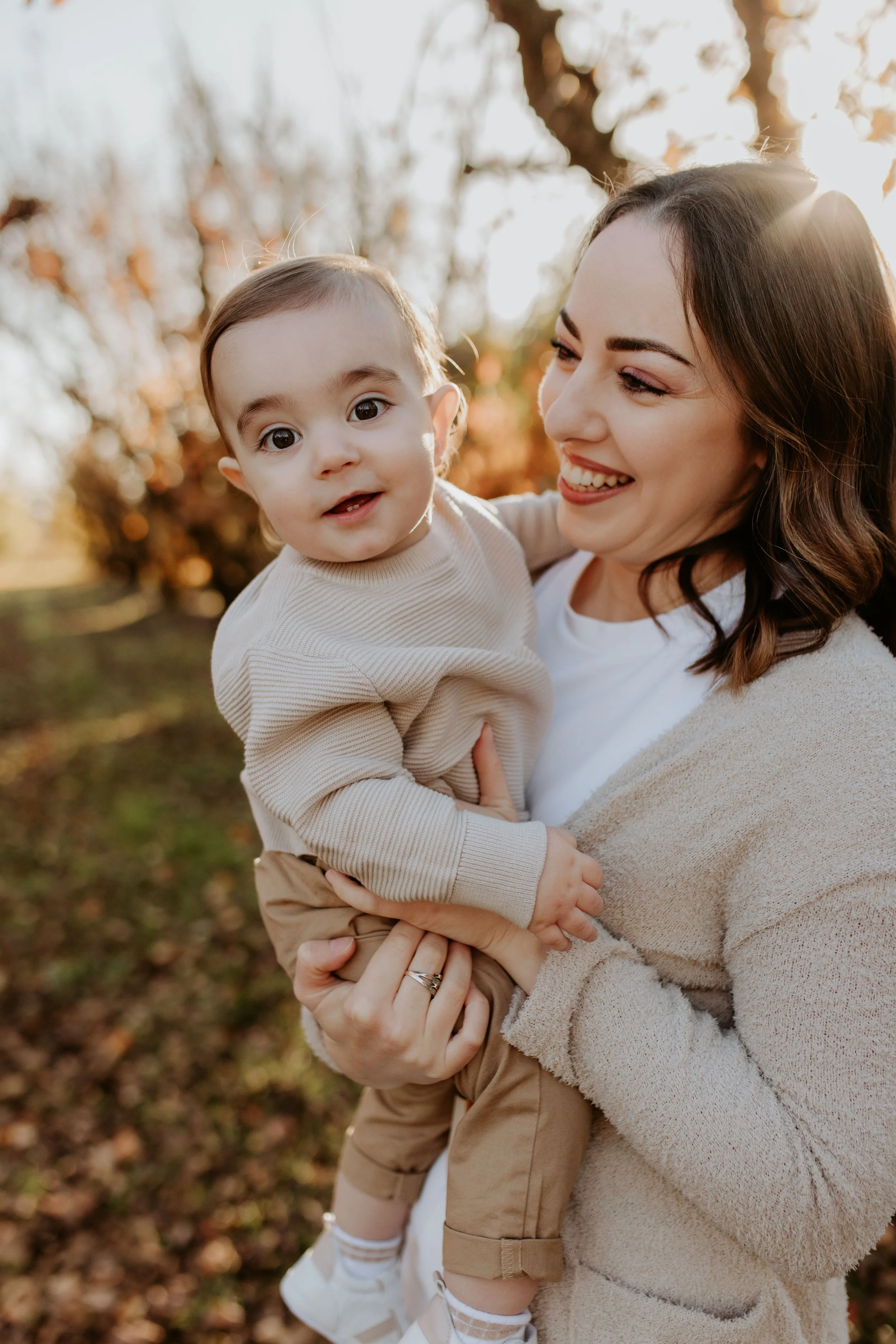 A smiling woman holding a young child outdoors in the fall with autumn trees in the background.