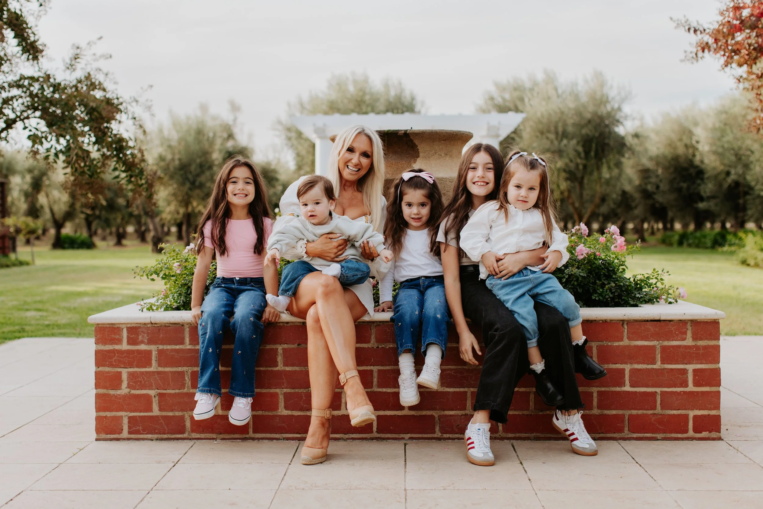 A woman with five young girls sitting on a brick ledge outdoors in a park, smiling, with trees and grass in the background.