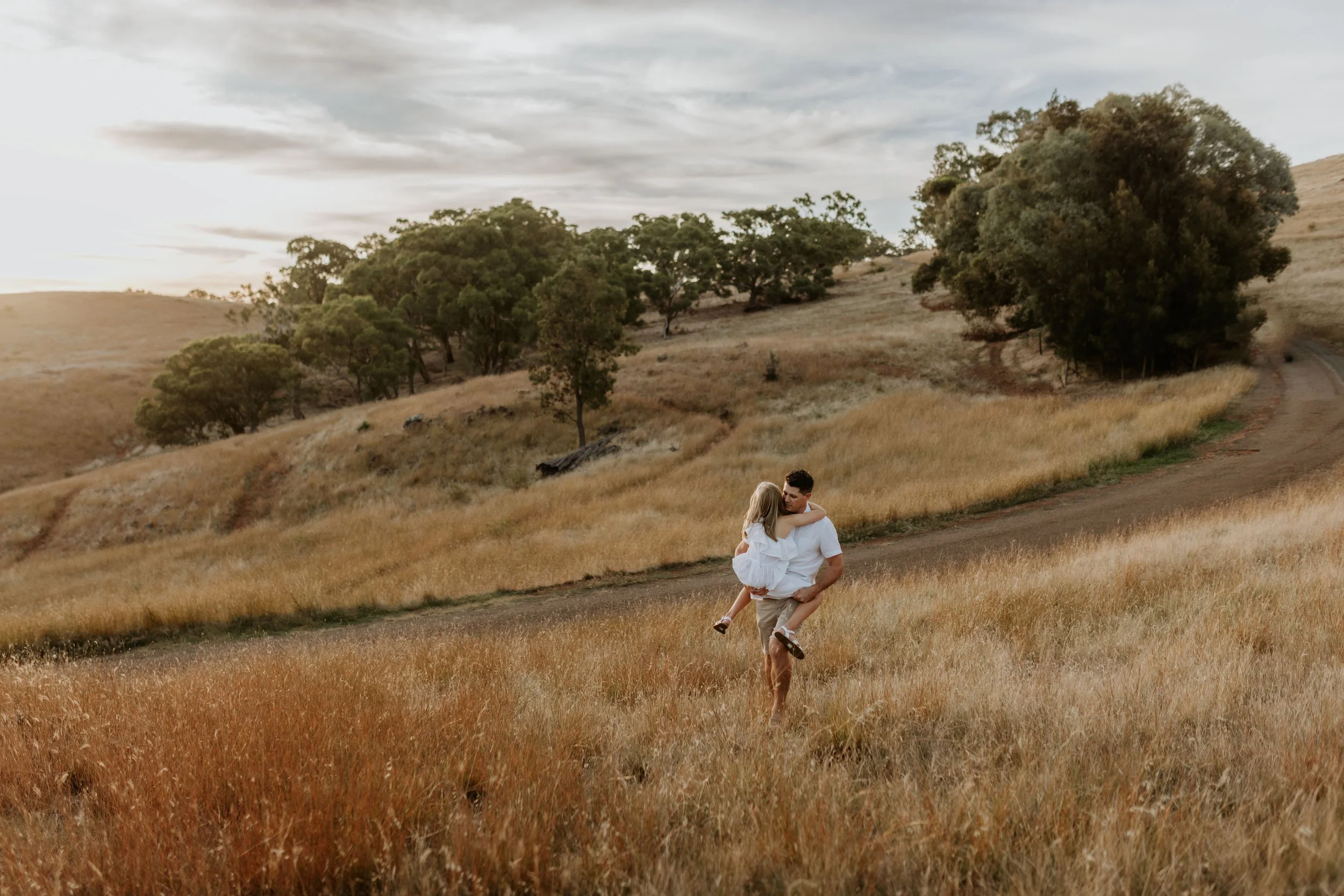 A man carrying his daughter in a white dress through a field of tall grass on a dirt road, with trees and hills in the background during sunset.