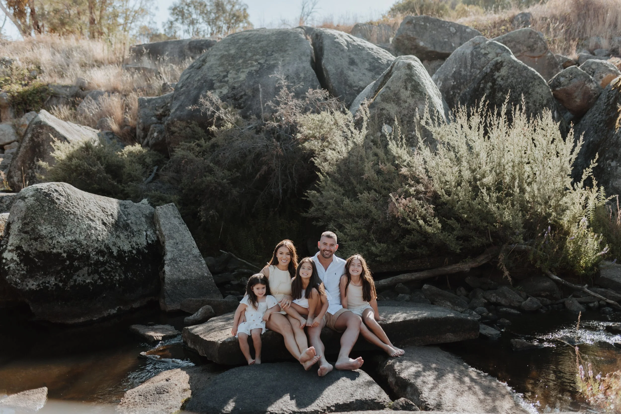 A family of five sitting on rocks by a stream in a natural outdoor setting with large rocks and green shrubs.