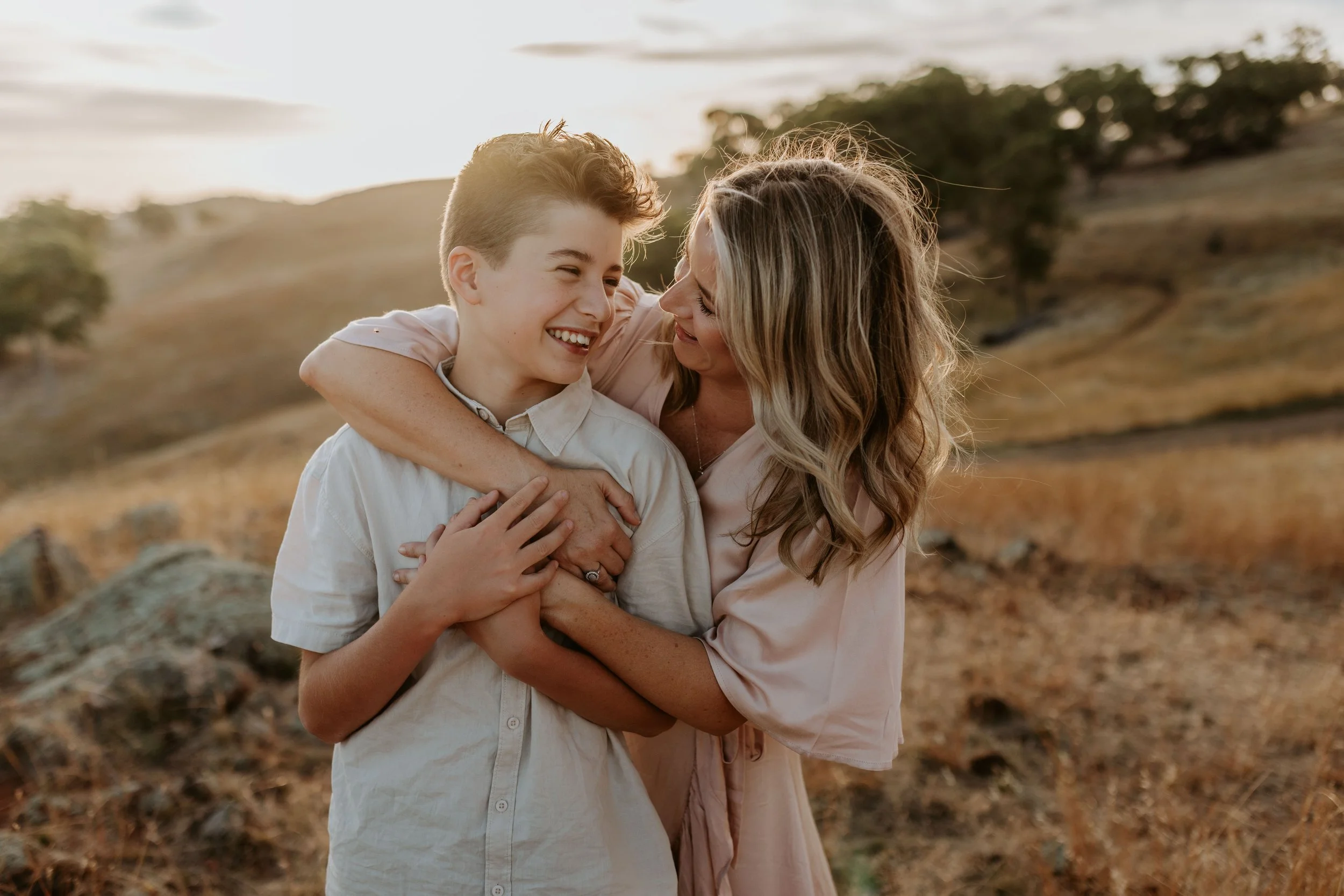 A woman and a young boy laughing and hugging in a grassy outdoor landscape at sunset.