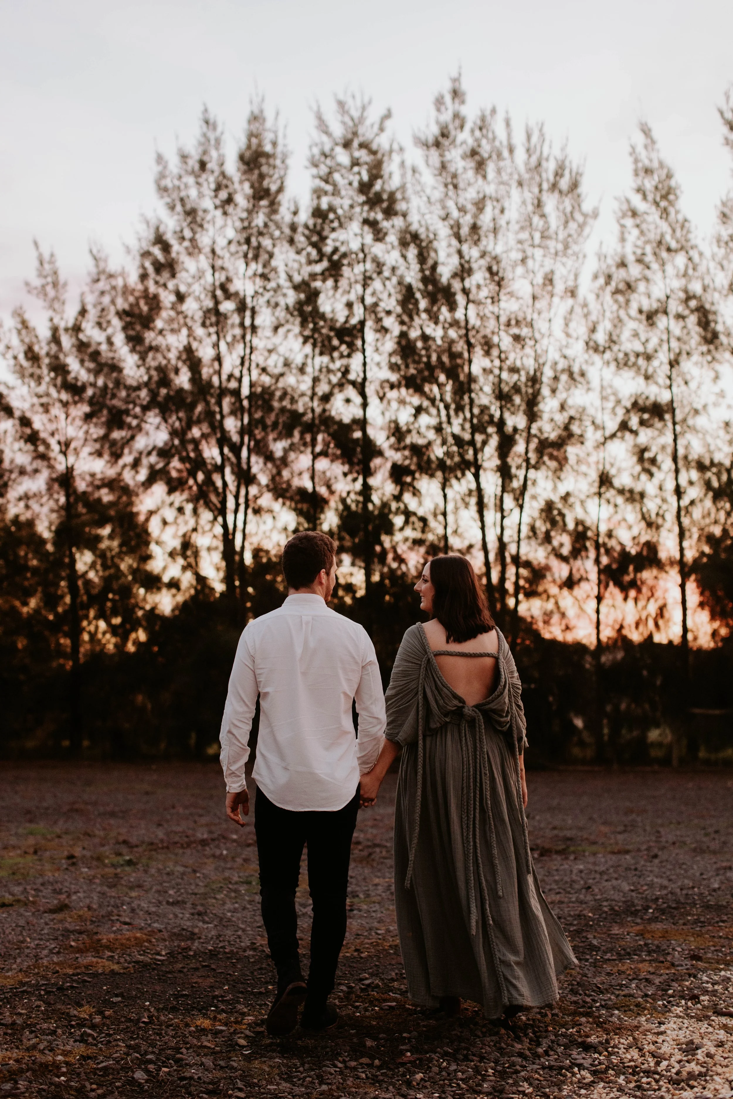 A couple holding hands and walking away in an outdoor setting at sunset, with trees in the background.