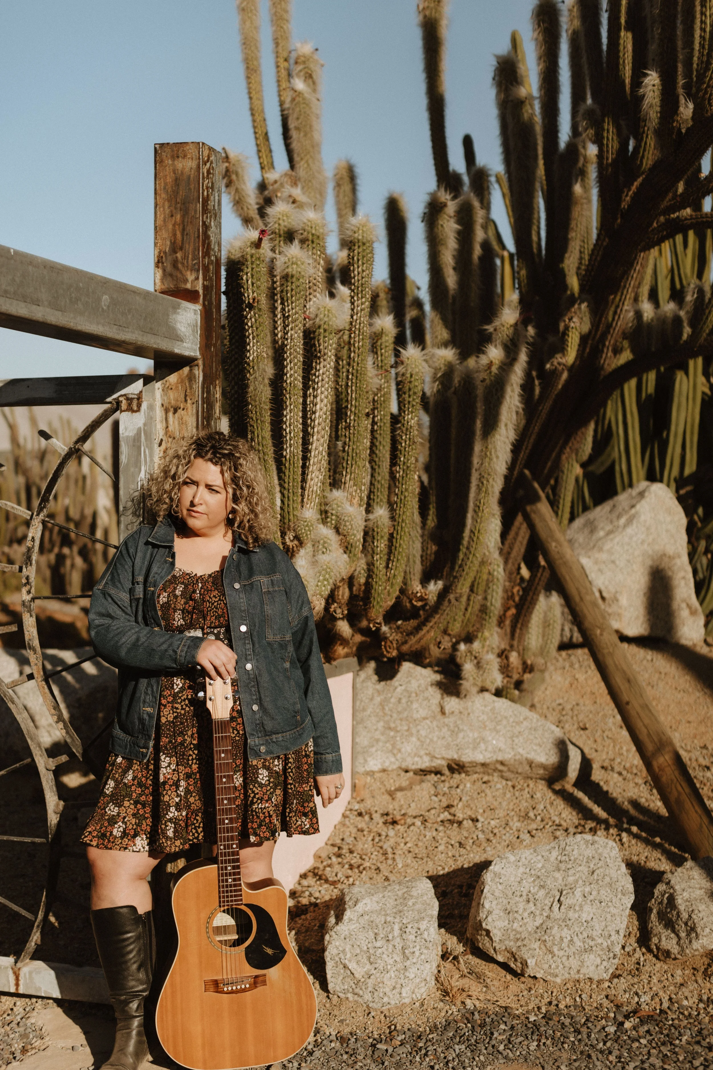 A woman with curly blonde hair wearing a denim jacket and a floral dress standing outdoors with a guitar, desert cacti, rocks, and a wooden structure in the background.