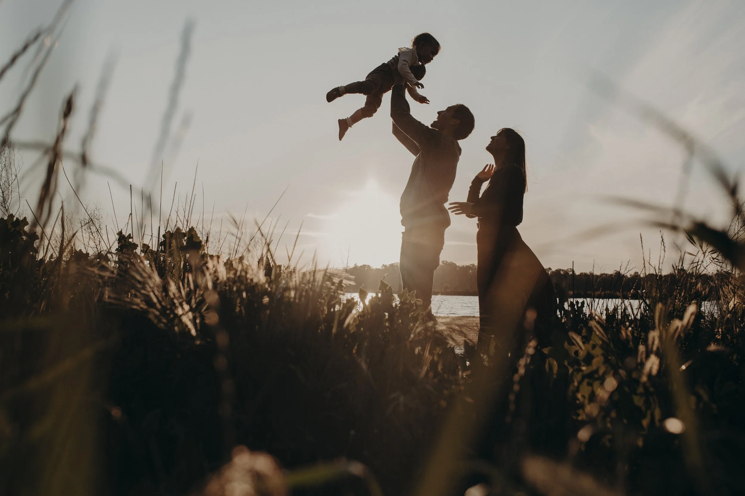 A family of three stands near a body of water during sunset. The father lifts the young daughter in the air while the mother looks on, all silhouetted against the setting sun.