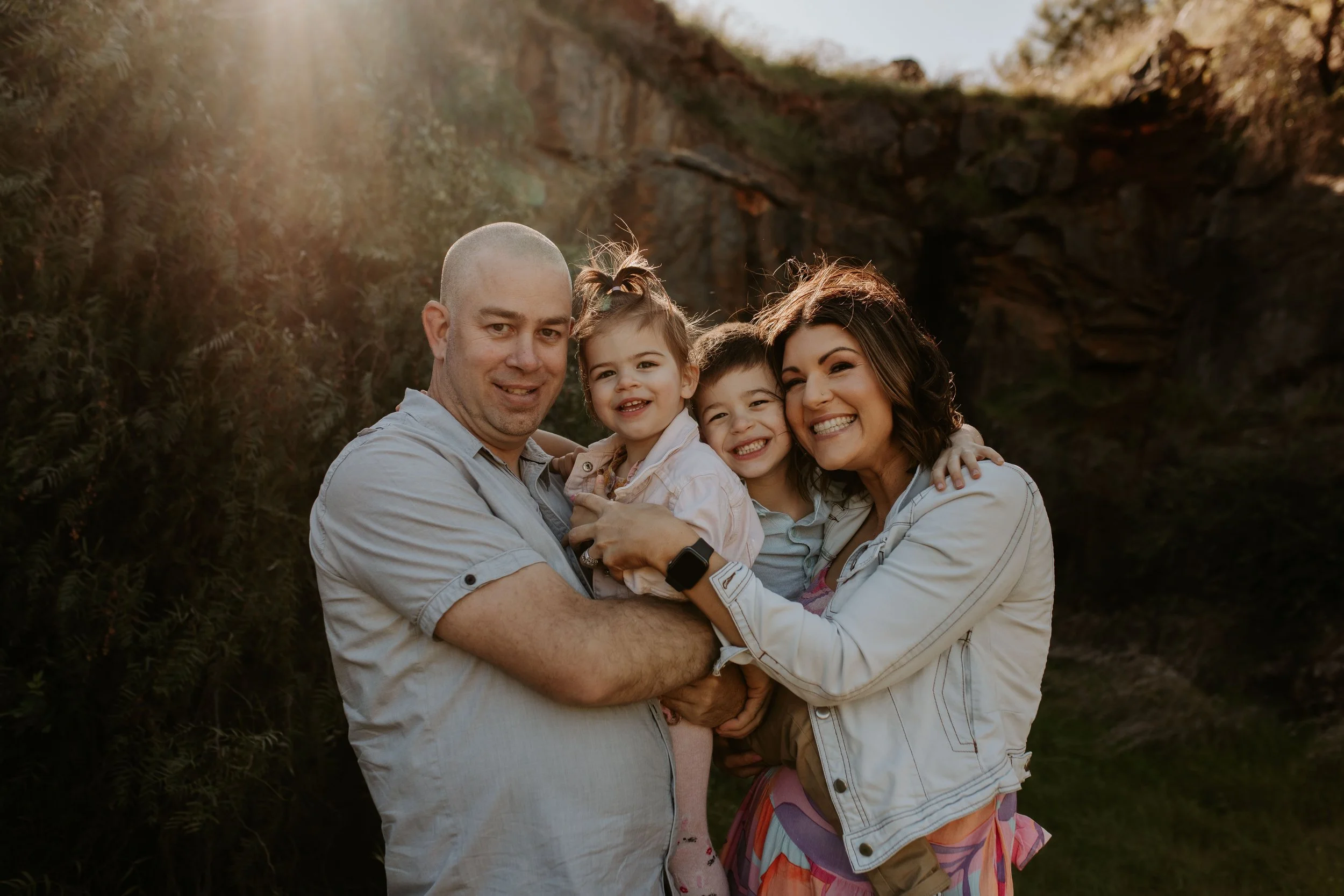 A happy family of four, with two children, outdoors during sunset, embracing and smiling at the camera.