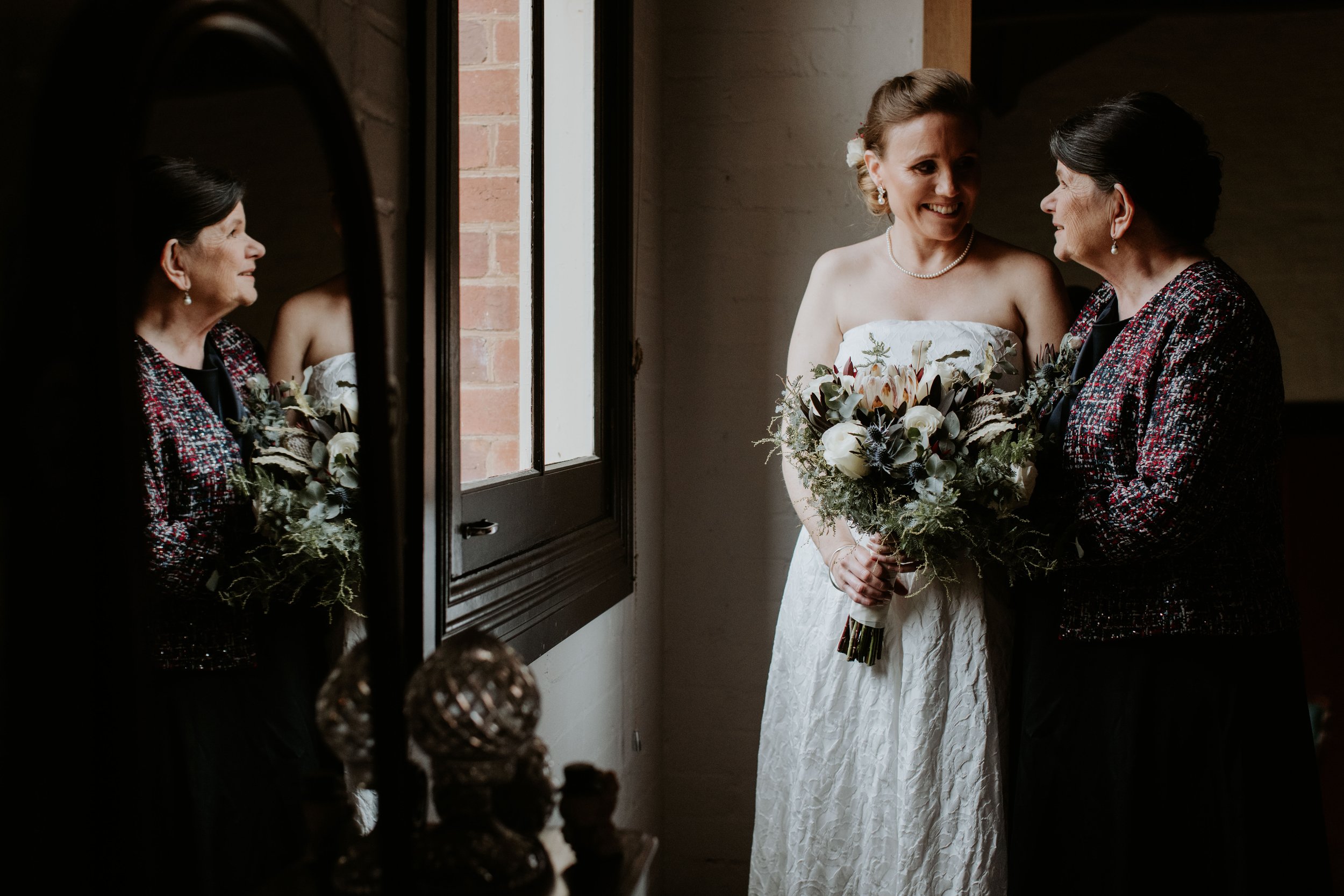 A bride holding a bouquet and smiling at an older woman, possibly her mother, inside a room with a window and brick wall.