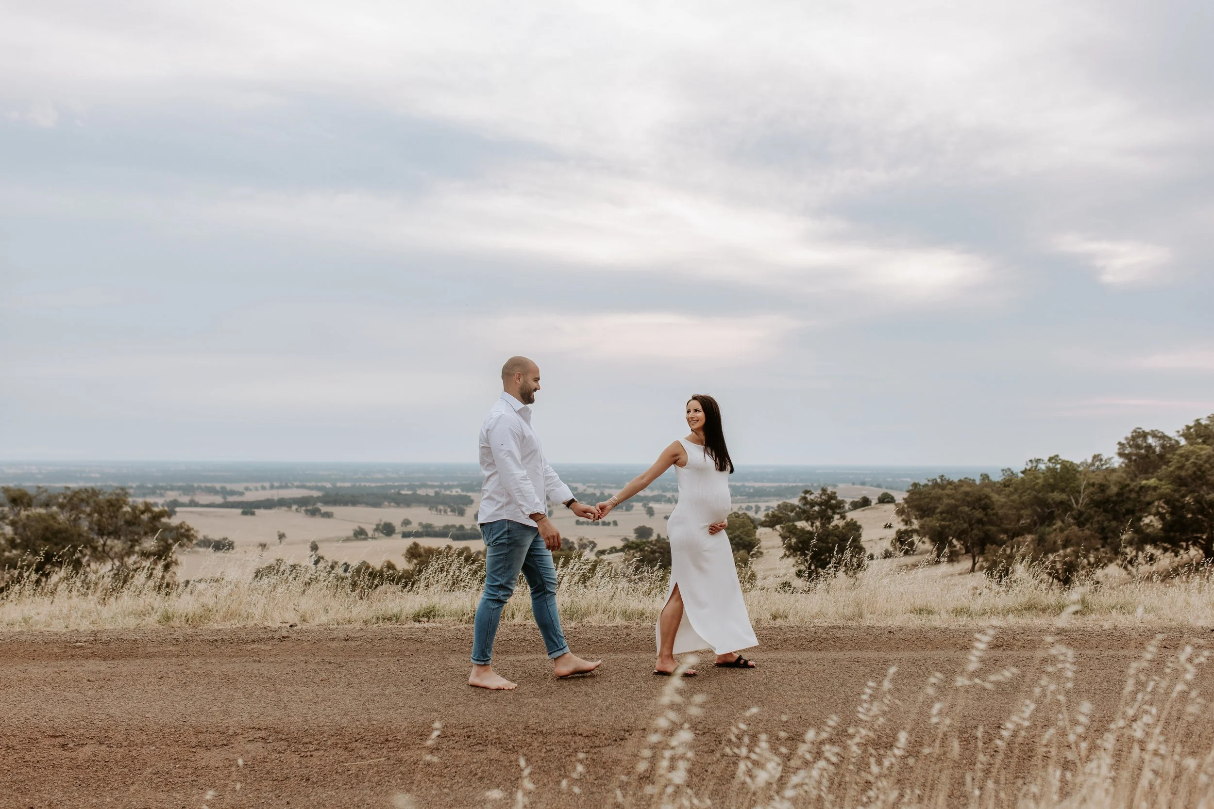 A pregnant woman in a white dress and a man in a white shirt and jeans holding hands and walking barefoot outdoors in a field with trees and cloudy sky in the background.