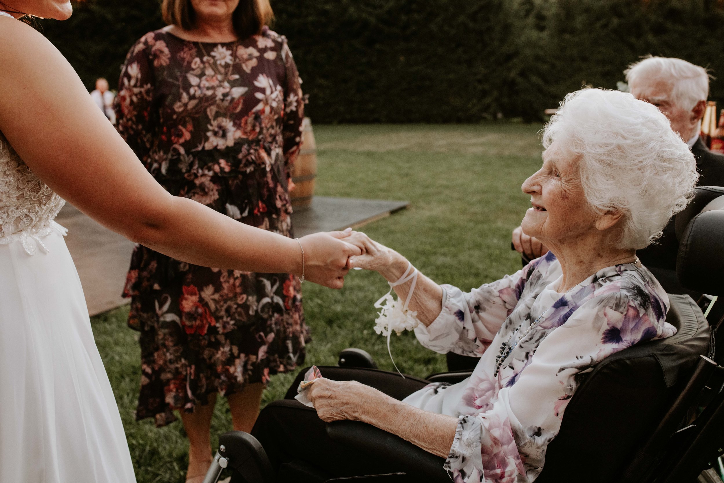 A bride holding hands with an elderly woman in a wheelchair at an outdoor wedding.