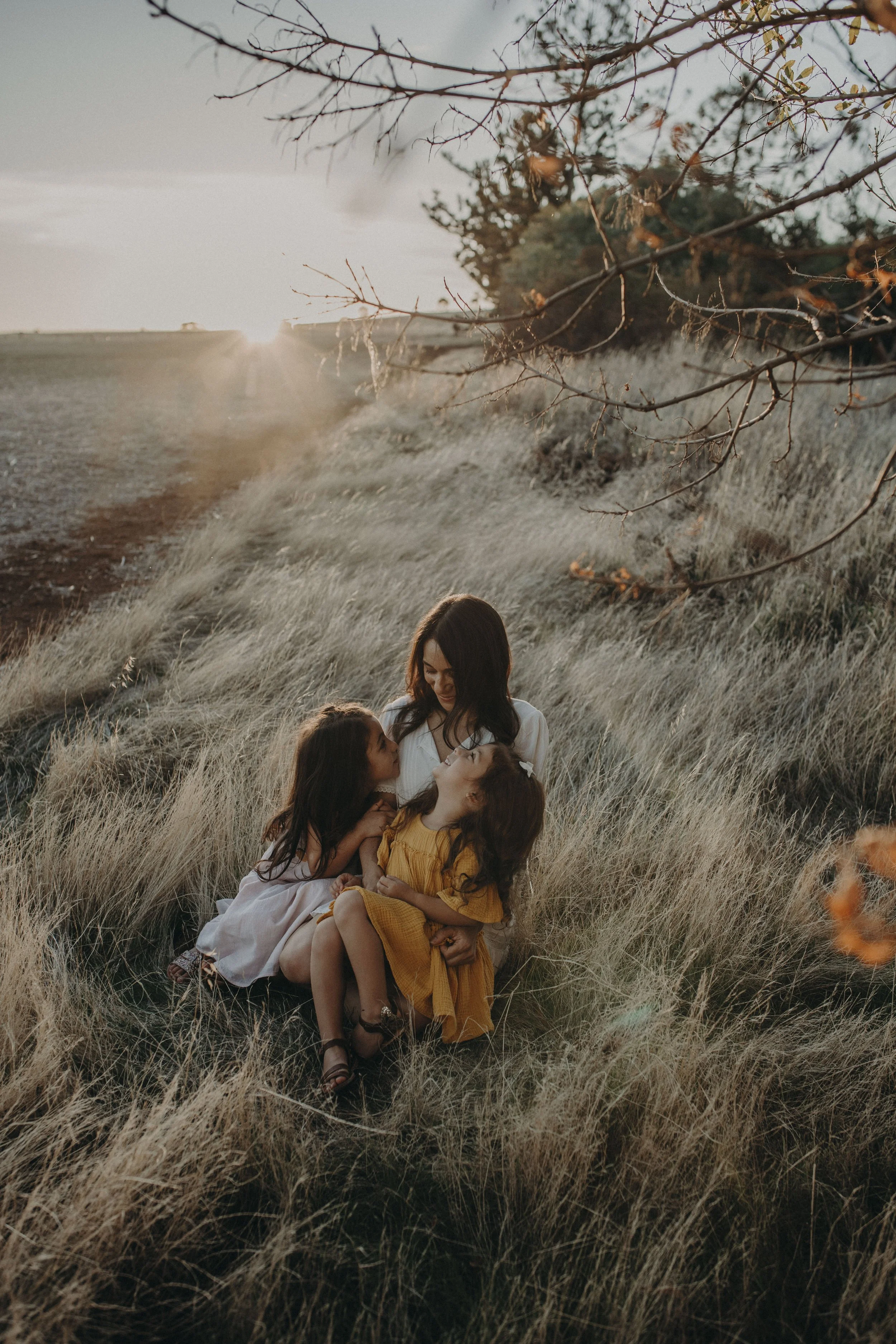 A woman with three young girls sitting in a grassy field during sunset, with trees and a clear sky in the background.