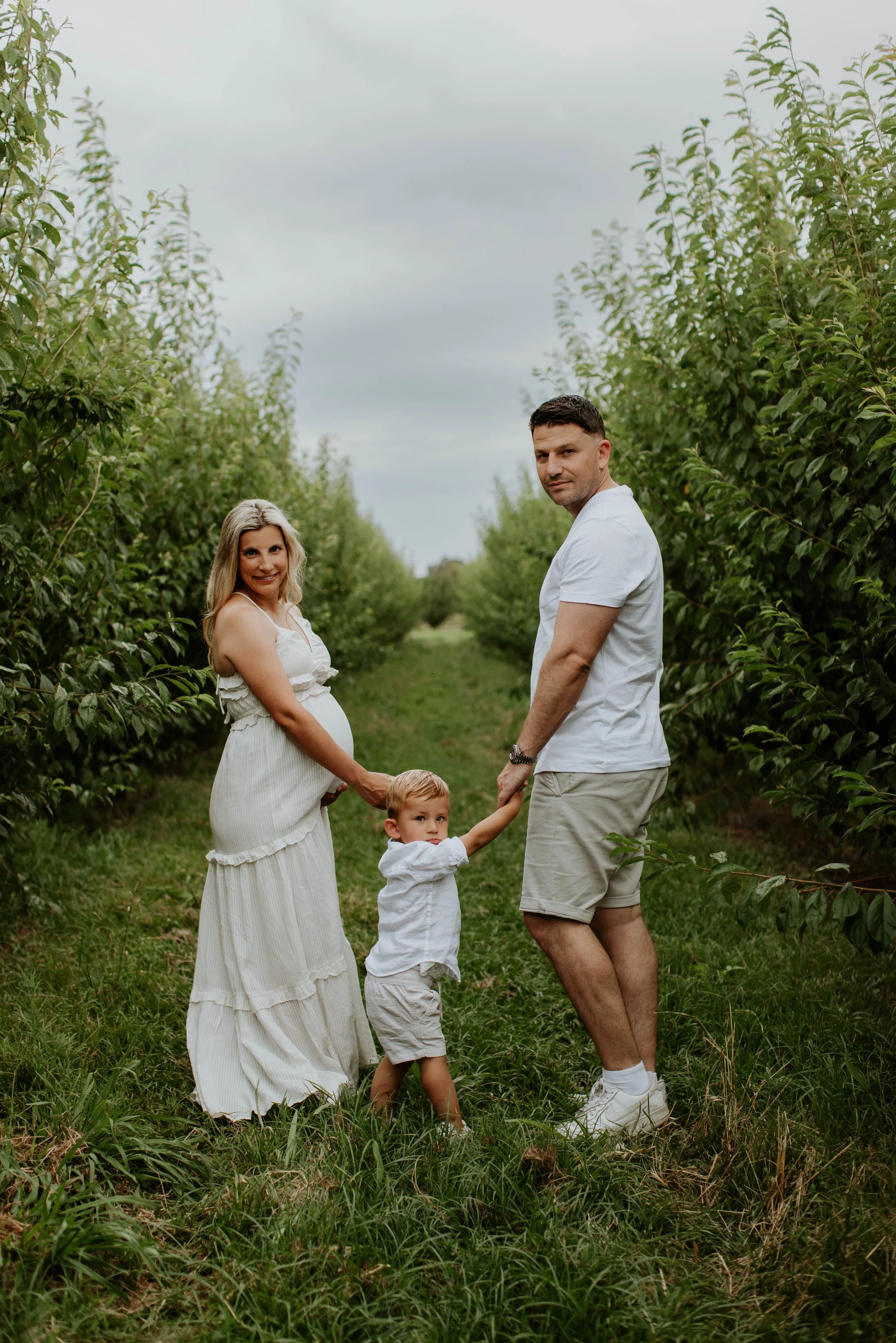 A family of three holding hands in an orchard, with a pregnant woman, a man, and a young boy. All are wearing light-colored clothing and standing on a grassy path among green trees.