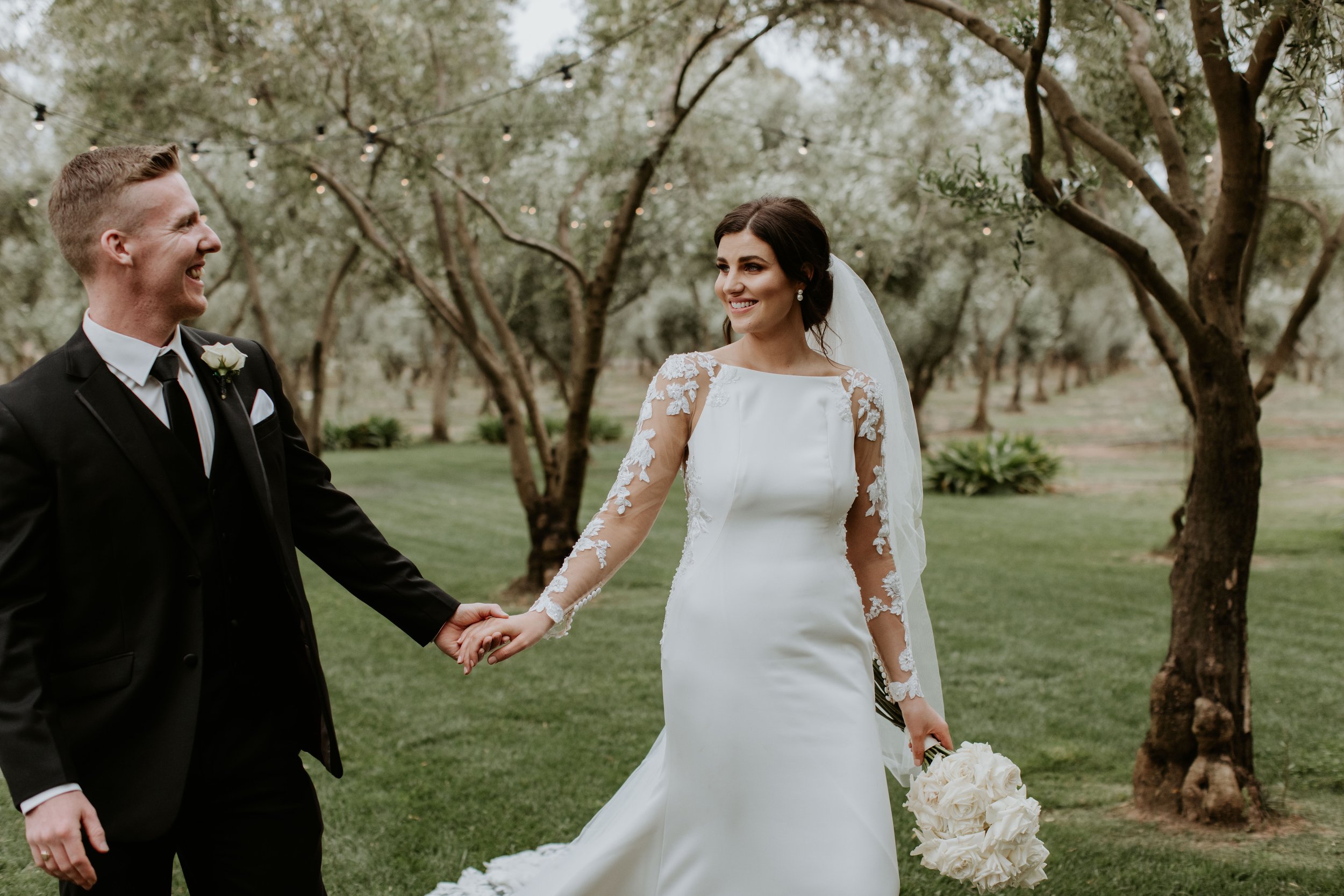 Bride and groom holding hands outdoors, smiling at each other, with trees and string lights in the background.