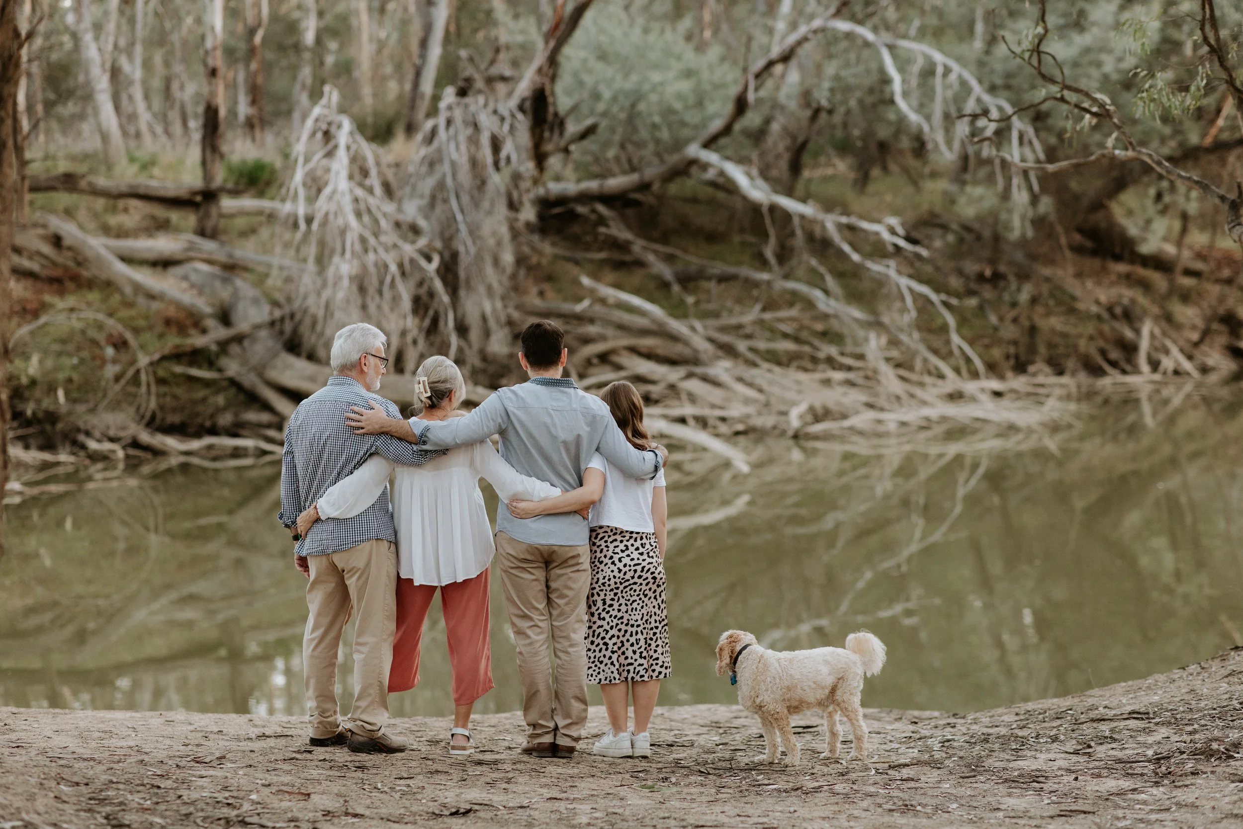 A family of five, including an elderly man, elderly woman, a man, a woman, and a dog, standing near a river, with the family embracing and looking across the water at a large fallen tree on a forested riverbank.