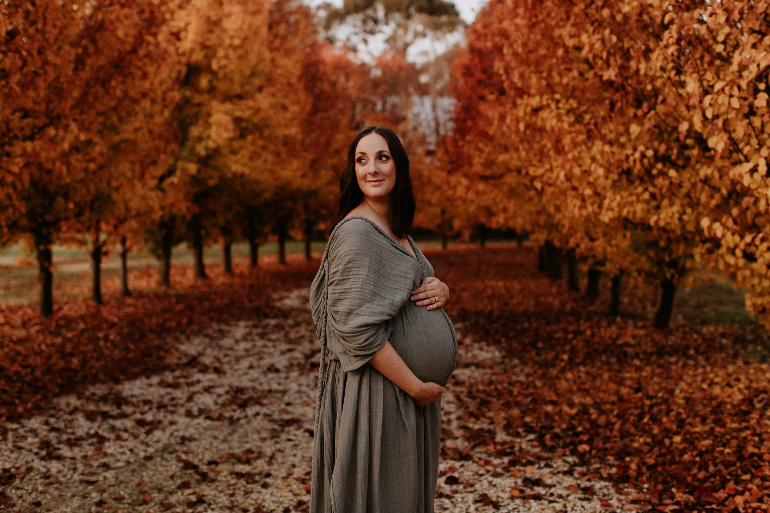 A pregnant woman standing on a leaf-covered path in an autumn-colored orchard, looking over her shoulder with a gentle expression.