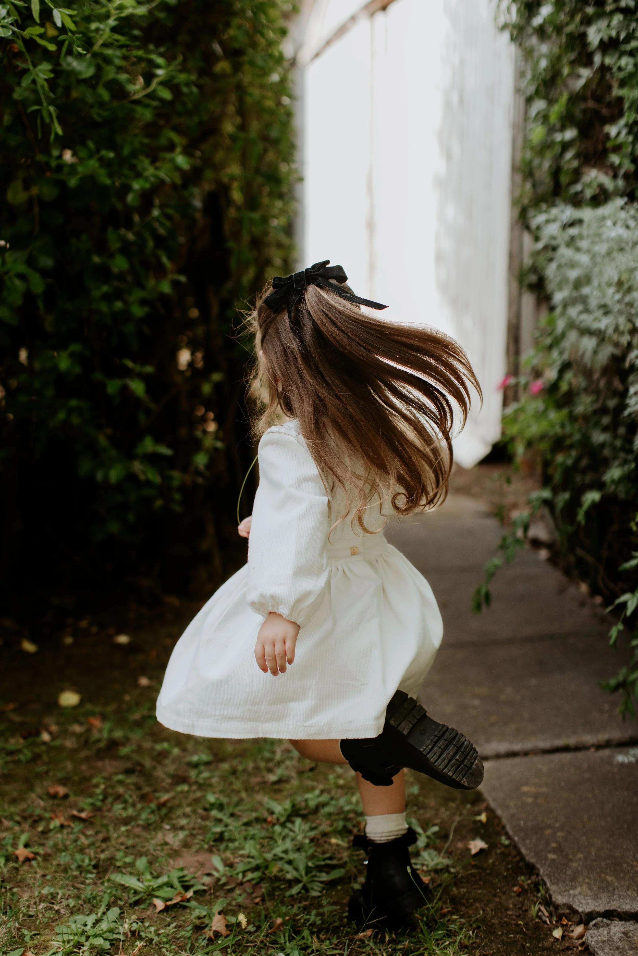 A young girl with long hair, wearing a white dress, black boots, and a black bow in her hair, spinning or twirling outdoors on a garden path surrounded by greenery.