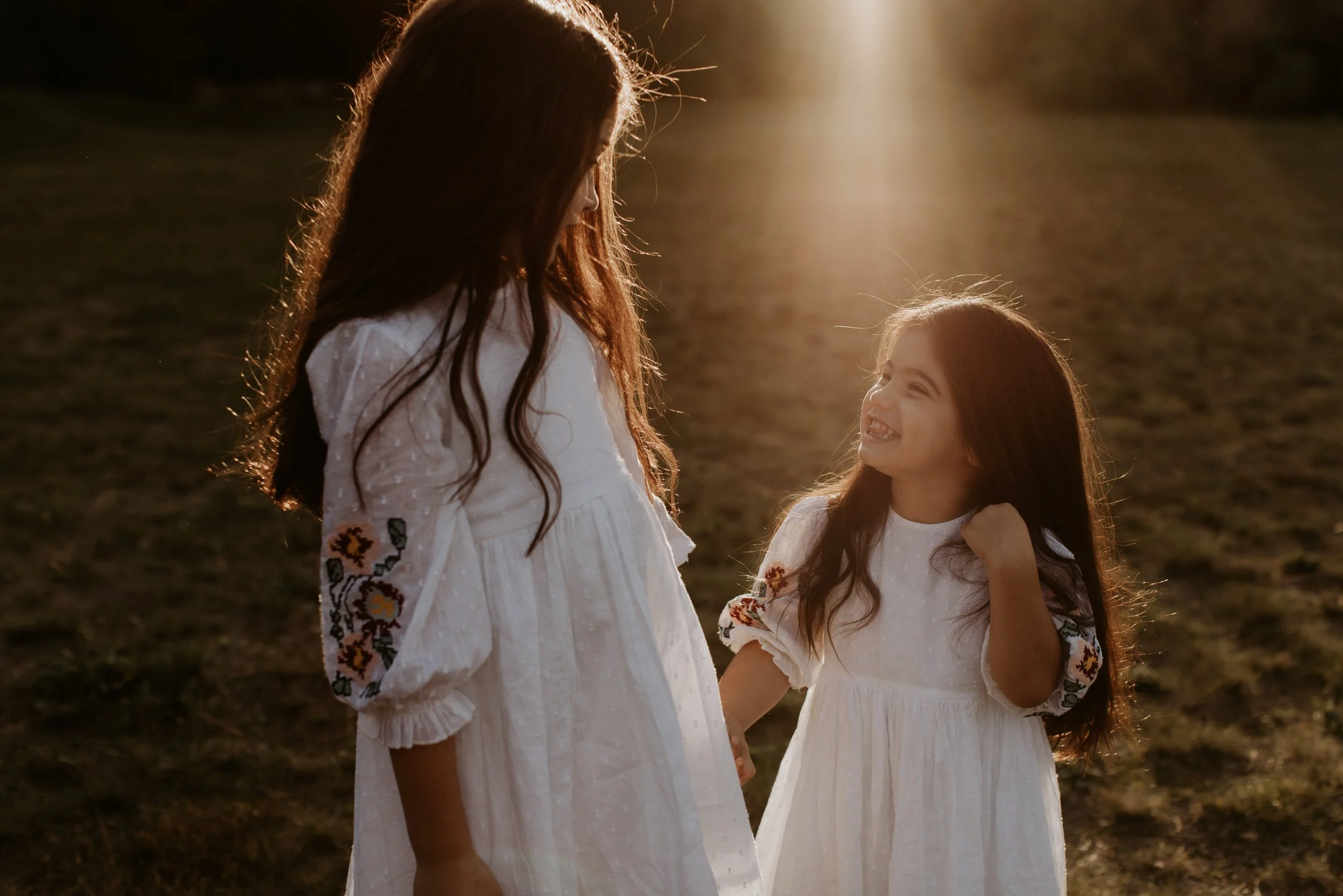Two girls with long brown hair and white dresses with embroidered sleeves. They are outdoors at sunset, smiling and holding hands, with a grassy field in the background.