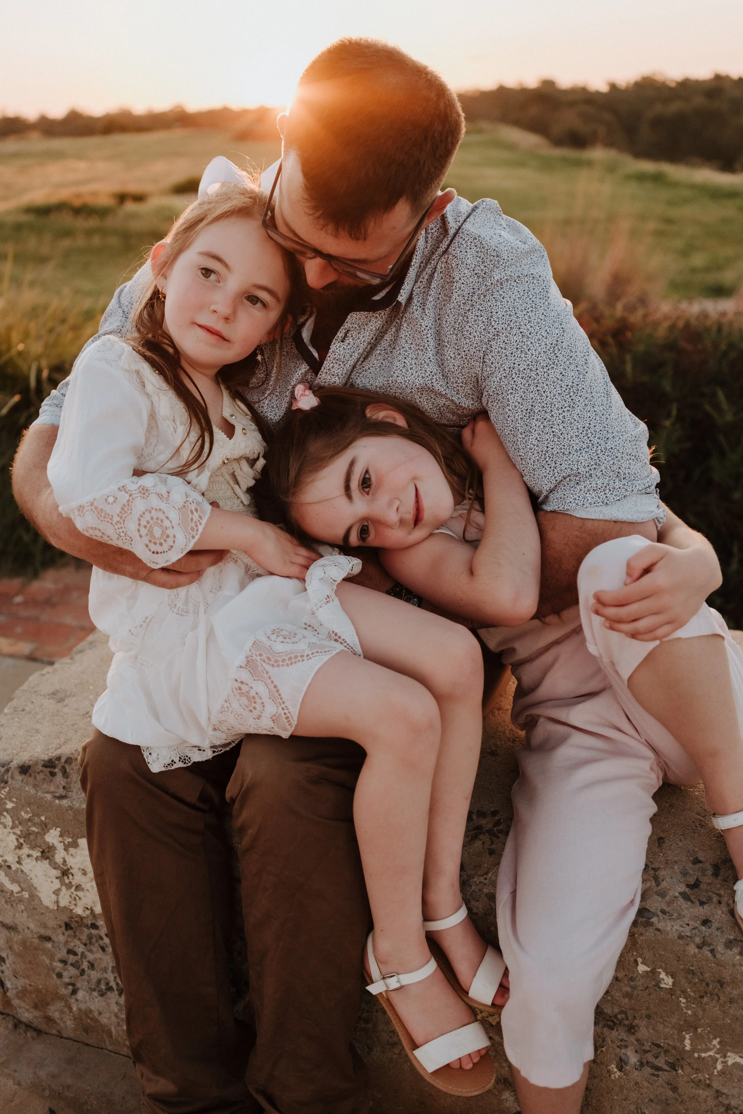 A man with two young girls in an outdoor setting during sunset, hugging and showing affection.