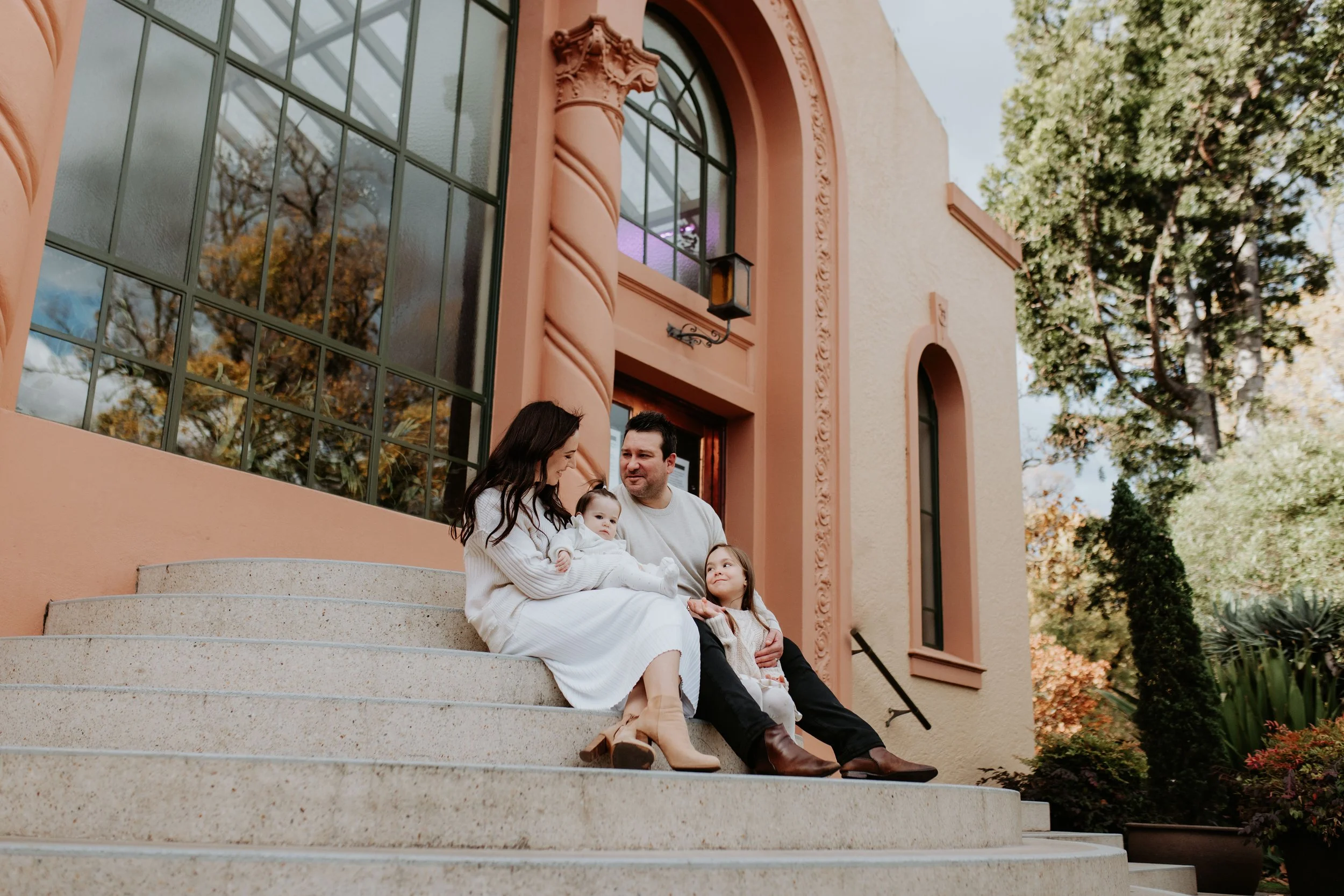 A family of four sitting on the steps of a peach-colored building, with two children and two adults, enjoying a moment together.