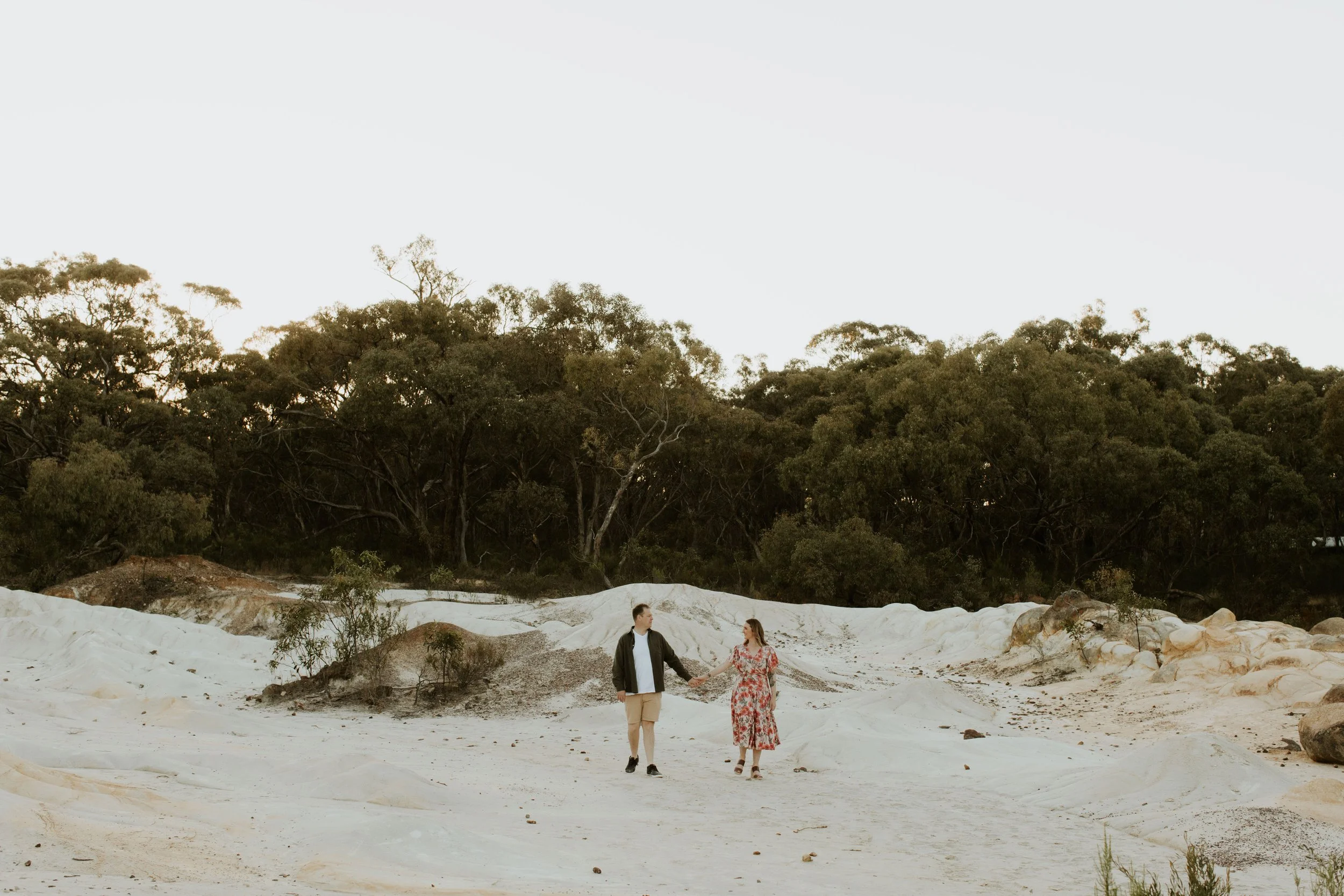 A couple holding hands walking across a sandy landscape with green trees in the background.