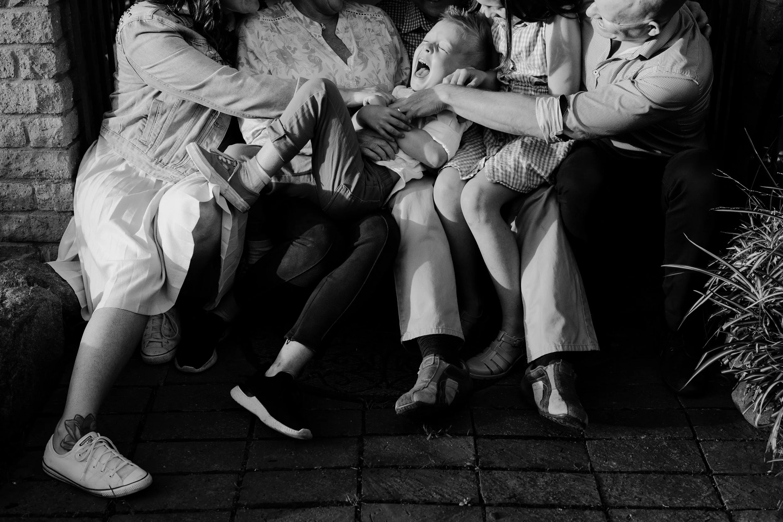 A black and white photo of children and adults playfully squeezing a boy who appears to be crying or yelling, on a brick patio with plants and a brick wall.