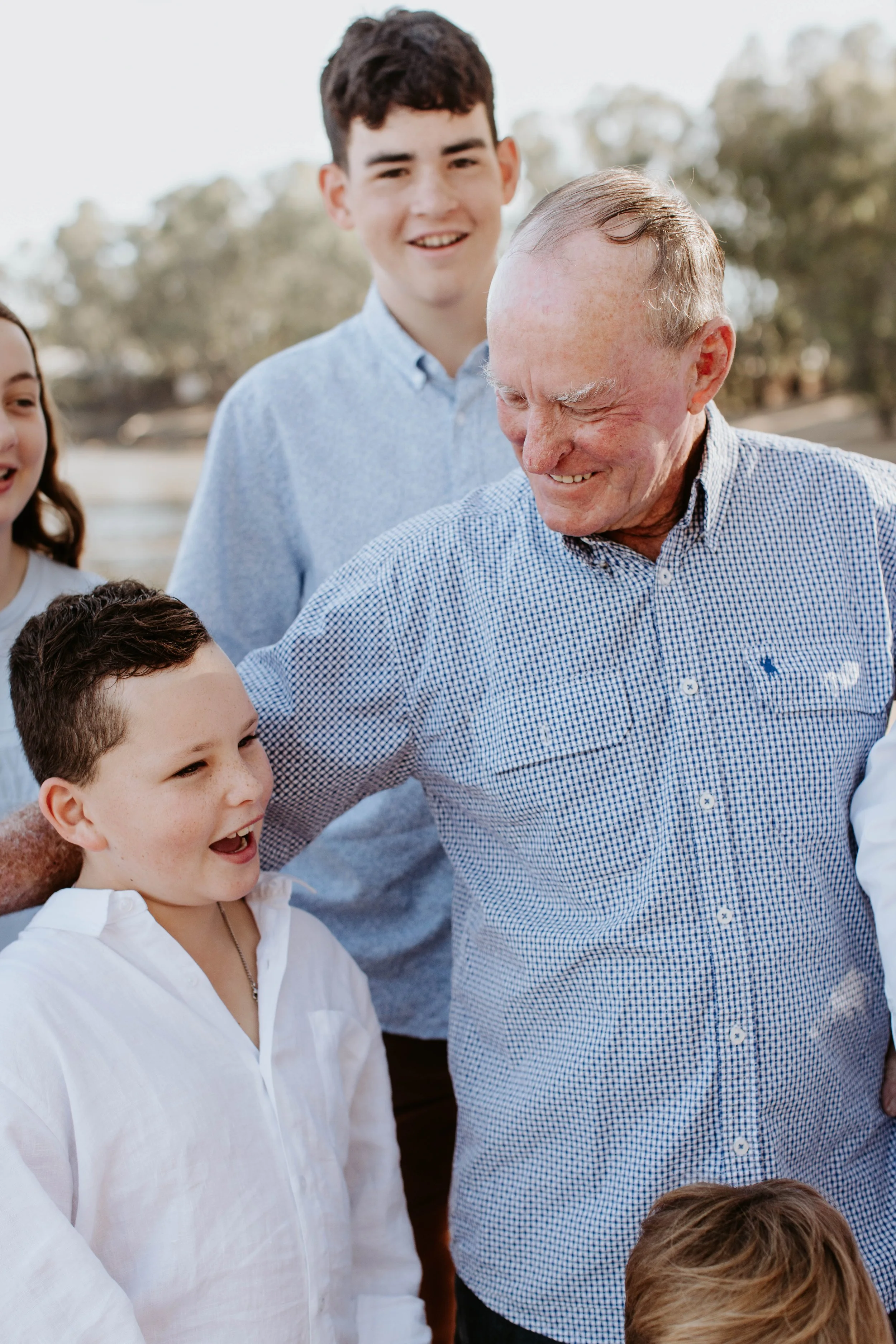 An elderly man smiling and talking to a young boy outdoors, with other children and trees in the background.