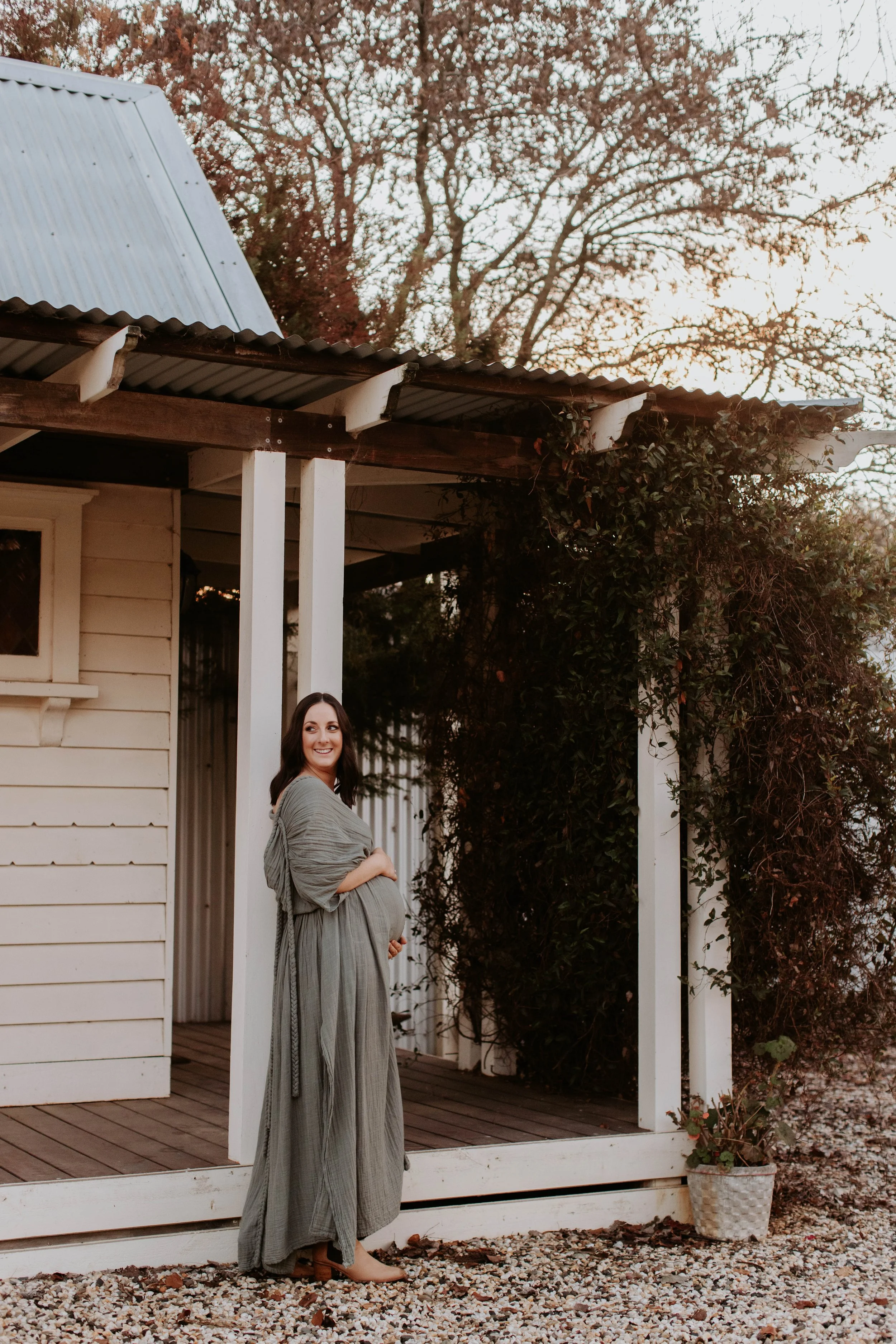 A pregnant woman standing outside a small wooden house, smiling and holding her belly, during sunset with trees in the background.