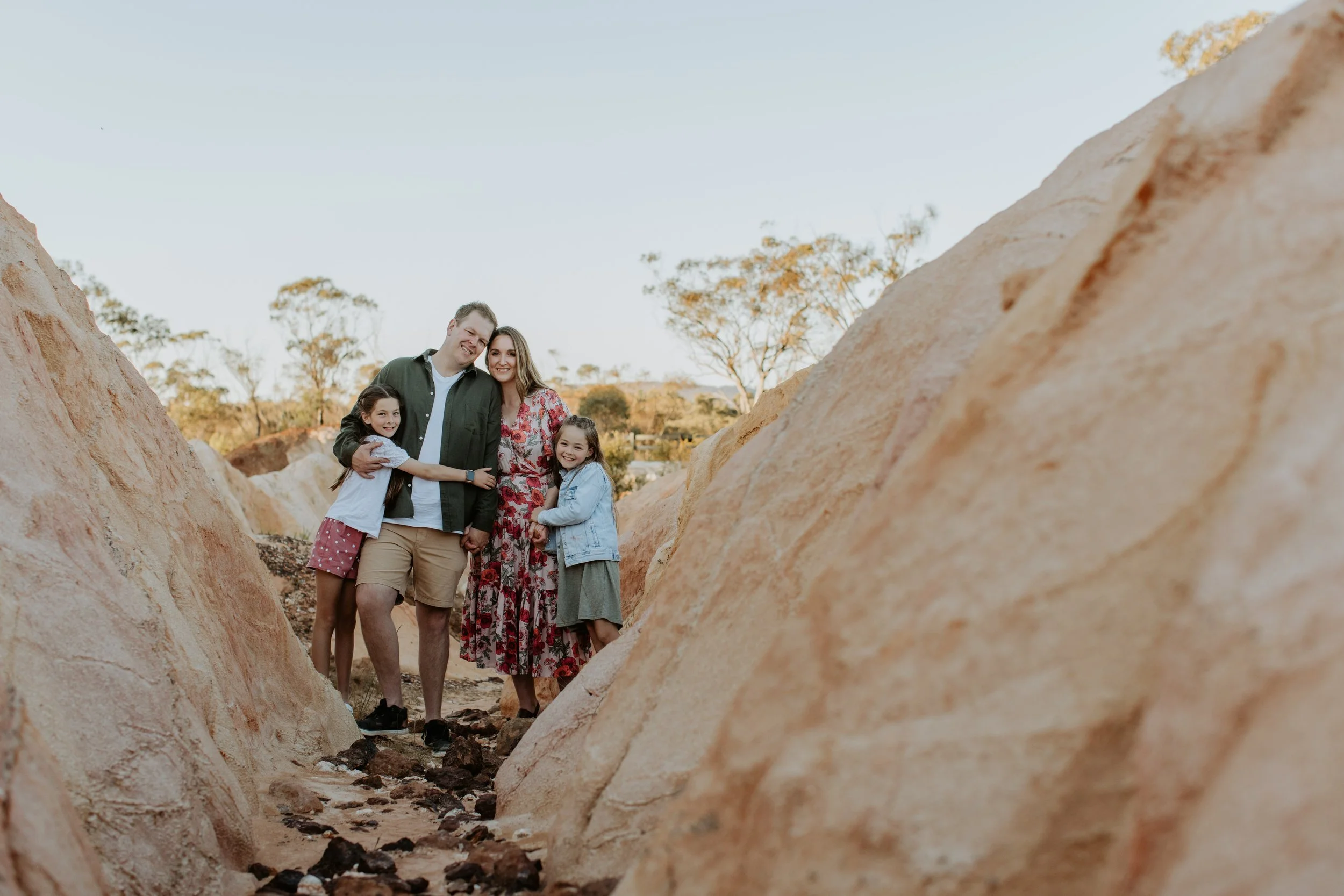 A family of four, two adults and two young girls, standing together outdoors between large rocks with trees and a clear sky in the background, smiling at the camera.