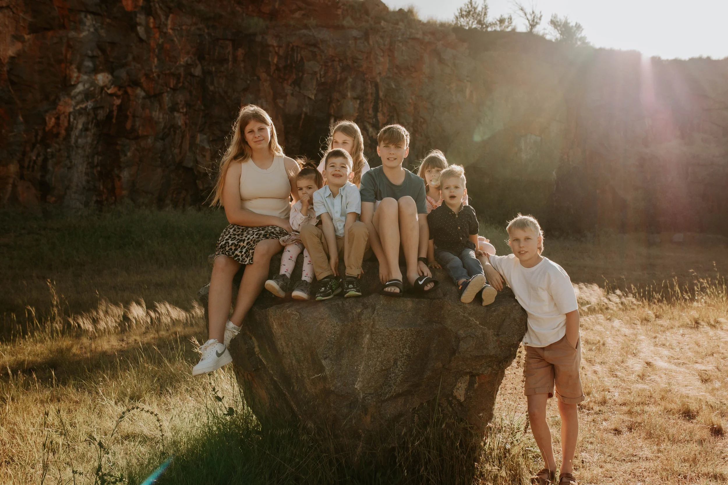 A group of children and a young woman sitting on and standing next to a large rock in a field with a rocky cliff in the background at sunset.