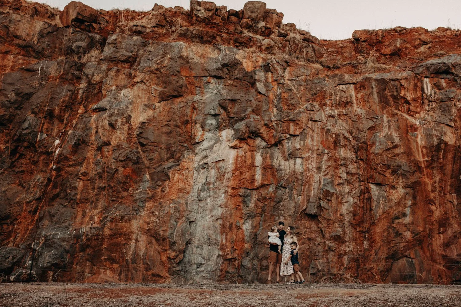 A family of four standing in front of a large, reddish-brown rocky cliff in a desert landscape during daytime.
