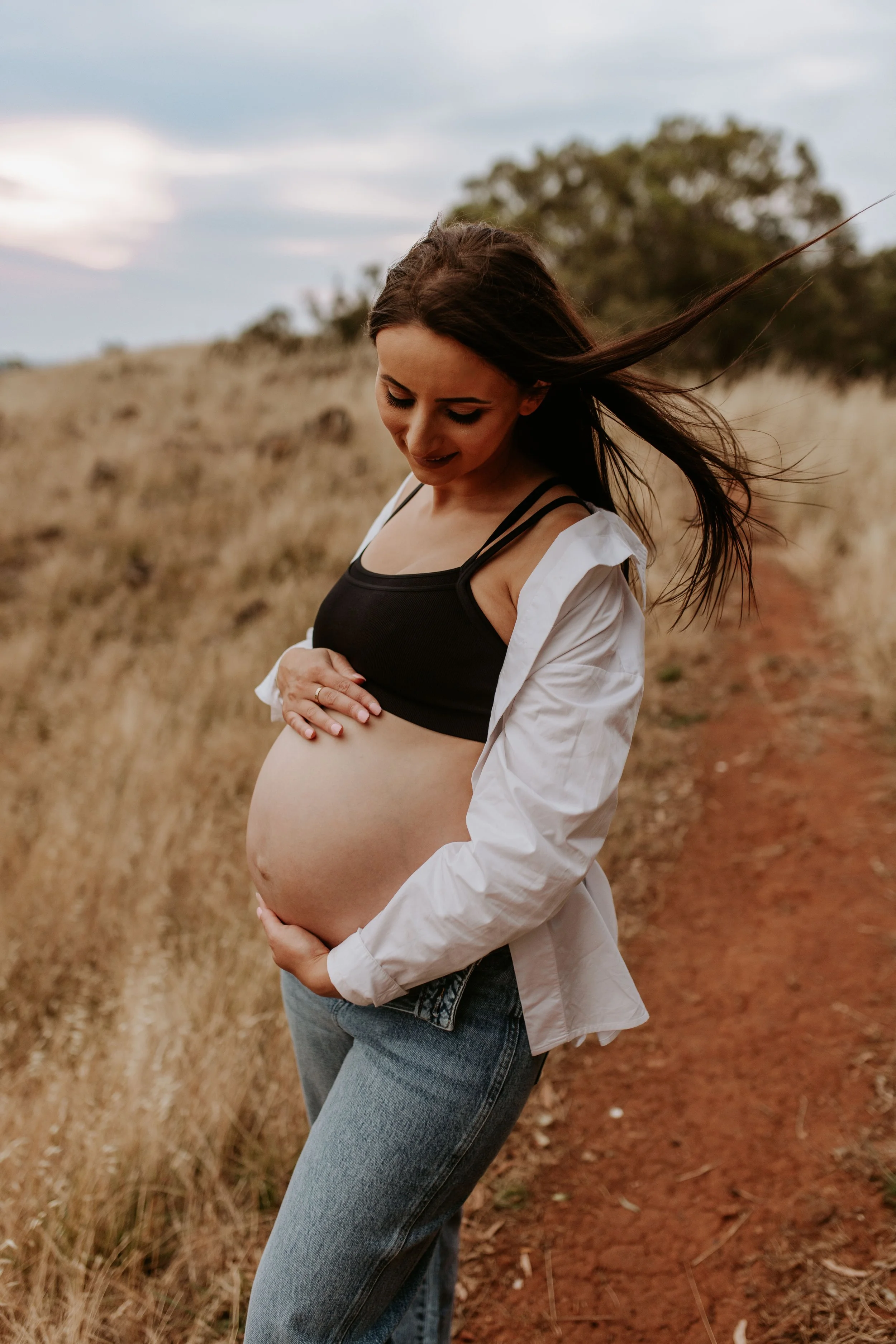 Pregnant woman standing on a dirt path in a grassy field, holding her belly, wearing a black top, open white shirt, and jeans, with long hair blowing in the wind.