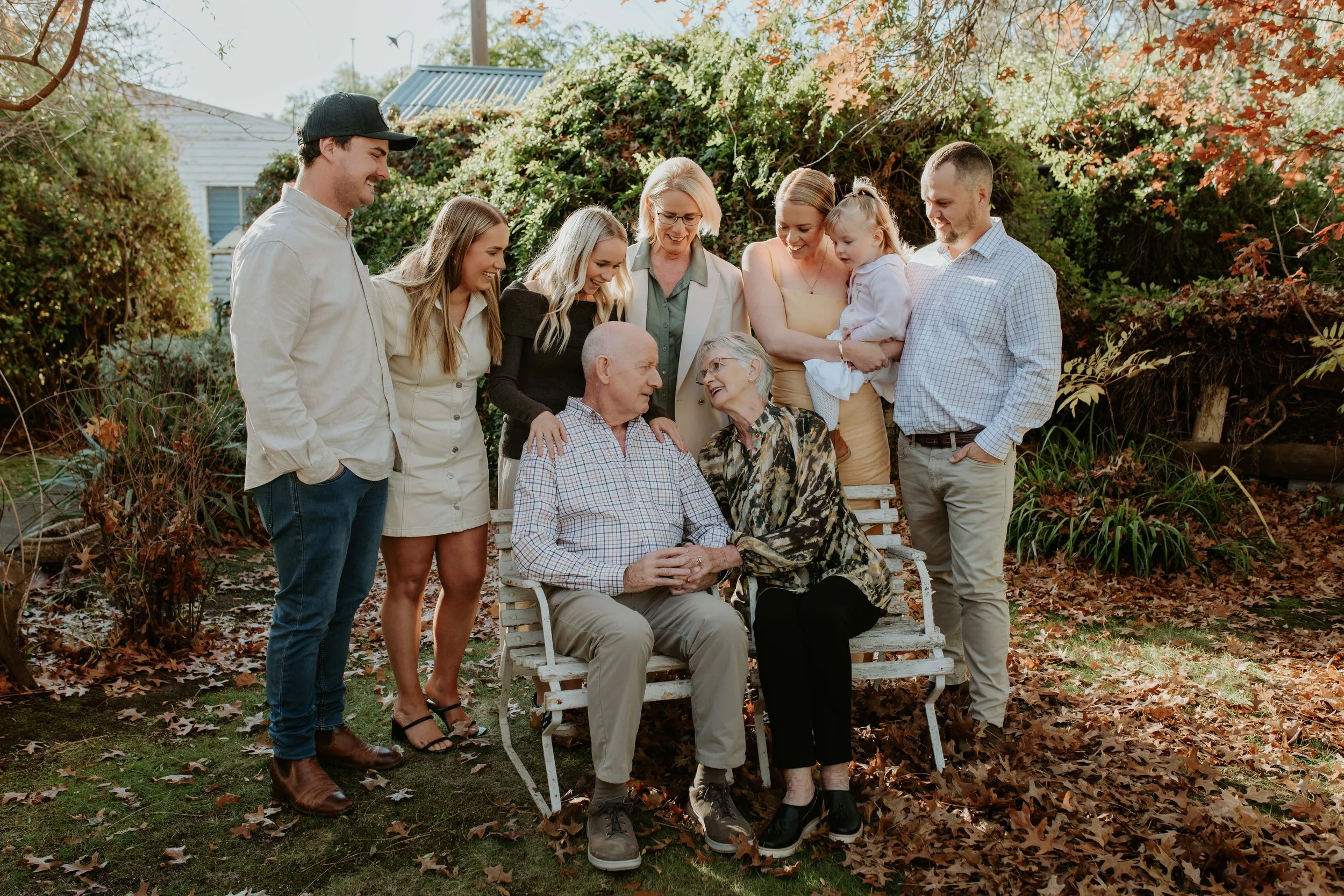 Family gathering outdoors in autumn, with elderly couple sitting on a bench surrounded by adult children and grandchildren, all smiling and engaging with each other.