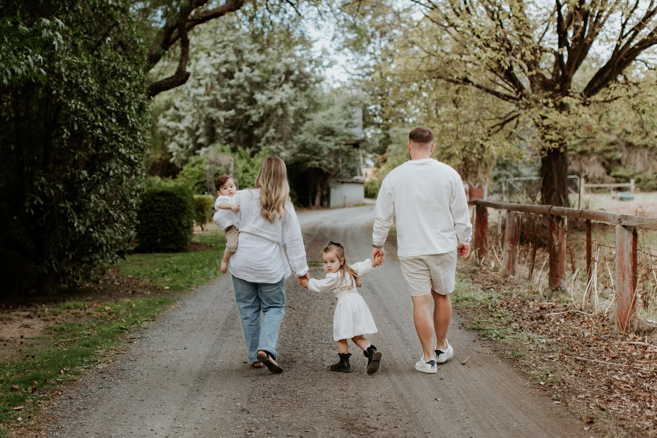A family of four walking hand-in-hand down a dirt path in a park surrounded by trees and greenery, enjoying a peaceful outdoor moment.